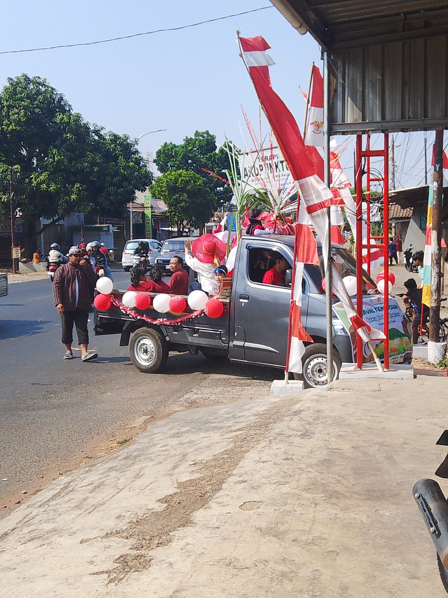 In the city of Kalinyamatan, Indonesia, a group of people are participating in a parade or celebration. A gray pickup truck decorated with red and white balloons and flags is the central focus. The truck is adorned with Indonesian flags and has a banner on the front that reads "KAVAL KEMBANG". Several people are visible on and around the truck. A man in a patterned shirt and dark pants is walking next to the truck. Inside the truck, there are passengers wearing red shirts. The truck is on a paved road, and there are trees and buildings in the background. Other vehicles and people are also present in the scene, suggesting a public event. The weather appears to be clear and sunny.