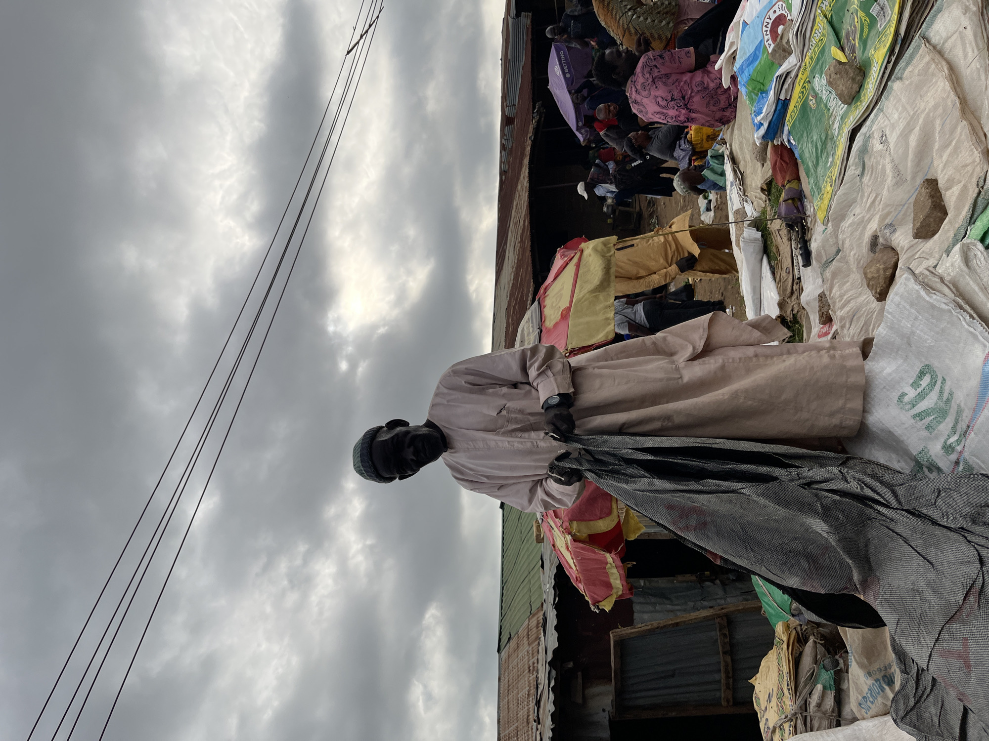 A man stands in a bustling outdoor market, holding a black mesh bag. He is wearing a light-colored robe and a knitted cap. In the background, other people are visible, along with colorful umbrellas and various goods stacked on the ground, including large sacks. The scene suggests a busy marketplace with many vendors and shoppers. The sky is overcast with dramatic clouds, indicating potentially gloomy or stormy weather. Overhead, several power lines crisscross the frame.
The image is oriented vertically. The overall atmosphere is one of lively commerce in a densely populated area. There is no specific indication of the time of day, but the lighting suggests it is daytime. The location cues point to Gura Top, Nigeria, a place known for its vibrant markets. The man's expression is neutral, and the general emotion conveyed is one of everyday activity.
Visible text includes "KG" on one of the sacks, and possibly "SUPERIOR OIL" on another, although these are partially obscured. The presence of various colored cloths, umbrellas, and sacks suggests a wide array of goods being sold. The setting appears to be a somewhat makeshift or informal market, with corrugated metal structures visible in the background.