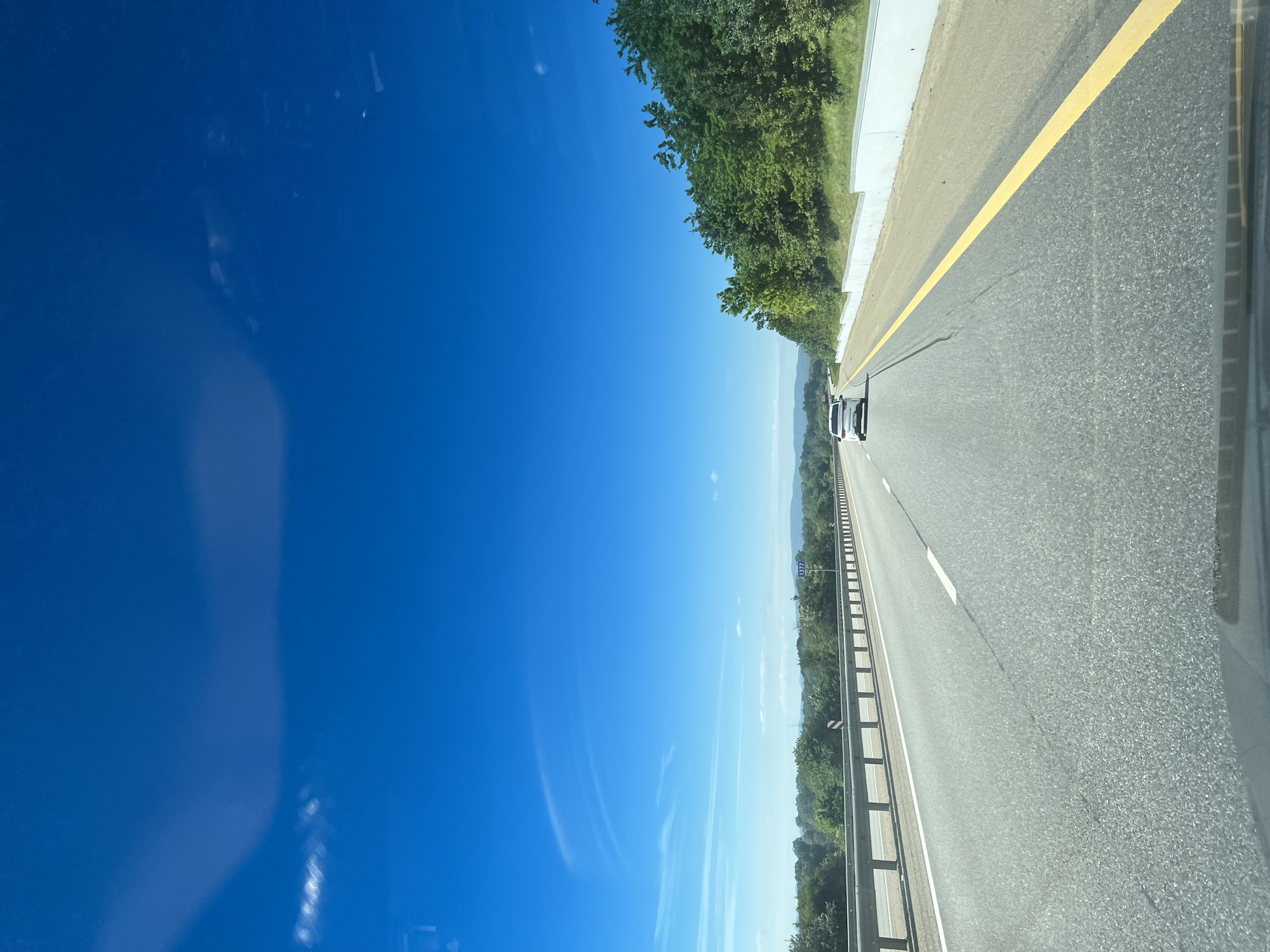 The image depicts a view from inside a vehicle on a highway. The highway is paved and has lane markings. Another vehicle is visible ahead. The surrounding environment includes trees and a clear blue sky. The location is Saratovskaya, Russia, as indicated by a road sign marked 1377.