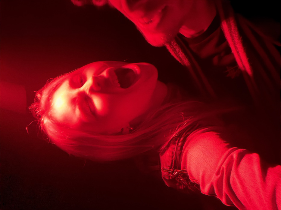 The image presents a close-up, chest-up shot of a female individual illuminated by intense red artificial light. The subject has light-colored hair and is captured mid-vocalization, with her mouth wide open, suggesting yelling or singing. Her eyes are partially closed. She is attired in a dark top beneath a denim jacket, which features a discernible zipper detail on the upper left arm area. Her left arm is extended into the foreground, while her right hand is partially visible on the right side of the frame, appearing to grip an object or clothing. The background is uniformly dark, indicative of an indoor, low-light environment, consistent with a nightclub, concert, or similar entertainment venue. No specific landmarks, architecture, or discernible environmental cues are present within the frame to identify a precise location within the city of Paris, France; the scene is confined to an interior setting.