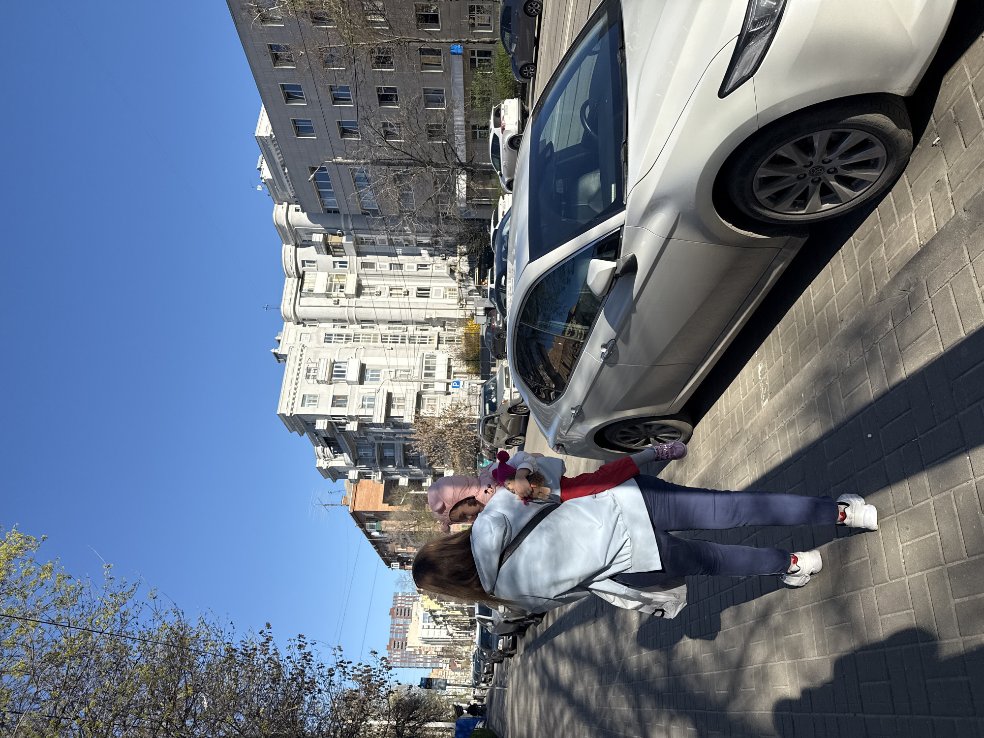 A daytime shot in Rostov-on-Don, Russia, features a woman in a light blue jacket, dark pants, and white sneakers carrying a small child in a pink hooded outfit and red pants. The child holds a stuffed animal. They stand on a brick sidewalk next to a white car parked on the street. Background buildings are mostly gray, multistory apartments, and trees line the street. The sky is clear and blue. Several other parked cars are visible down the street.