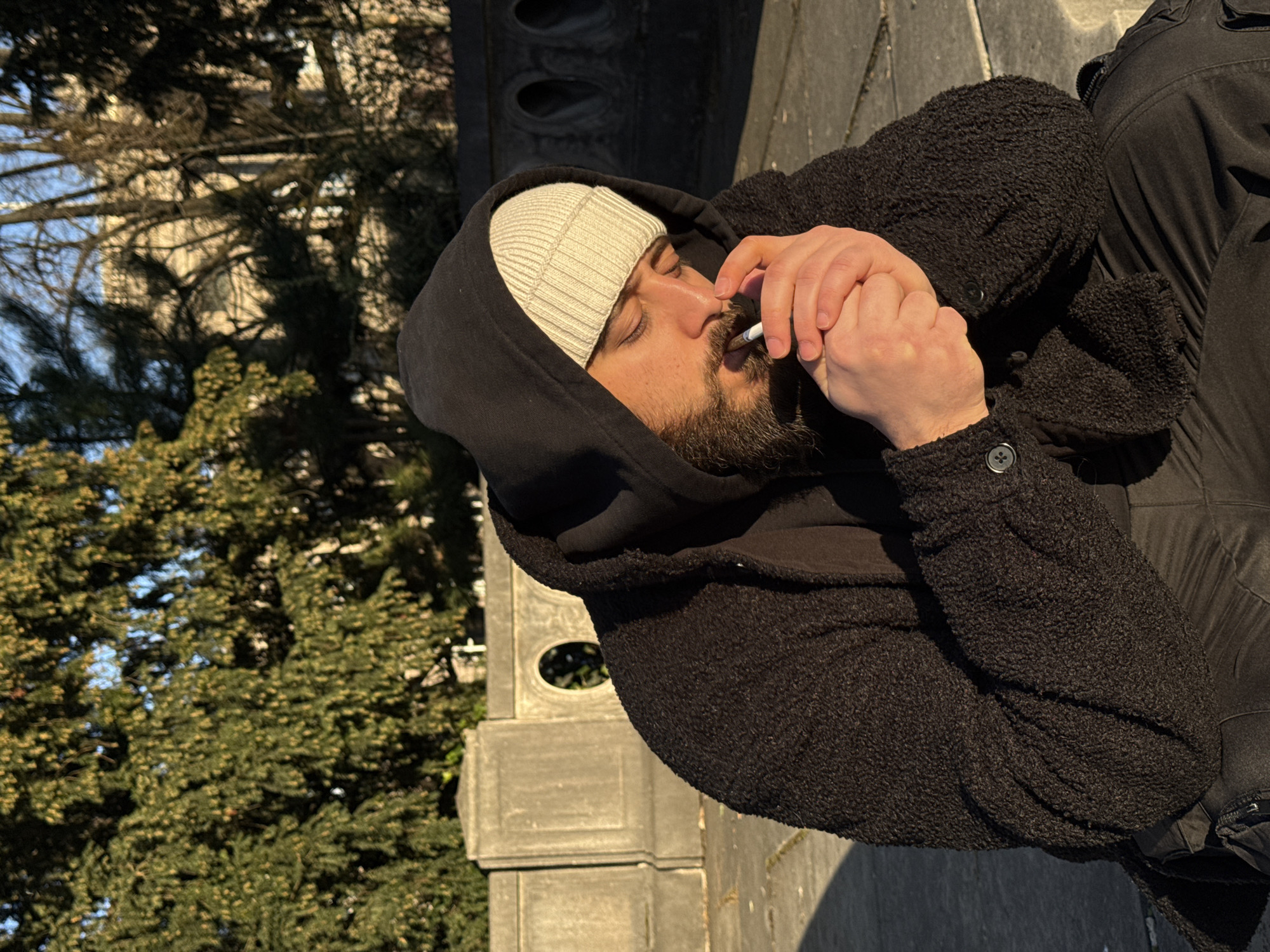 A male subject with a beard, wearing a cream knit beanie under a black hooded sweatshirt and a dark, textured jacket, is depicted outdoors in the Parc de l'Orangerie, Strasbourg, France. He is seated or crouched, actively lighting a cigarette held between his lips with a silver-colored lighter held in both hands. His gaze is directed downwards at the cigarette. Strong, low-angle sunlight illuminates the right side of his face and hands. In the background, a distinctive concrete or stone balustrade with circular openings is visible. Behind the balustrade are dense green evergreen trees, with bare deciduous trees and distant structures further beyond, under a clear sky. The ground surface in the foreground appears to be paved or stone.