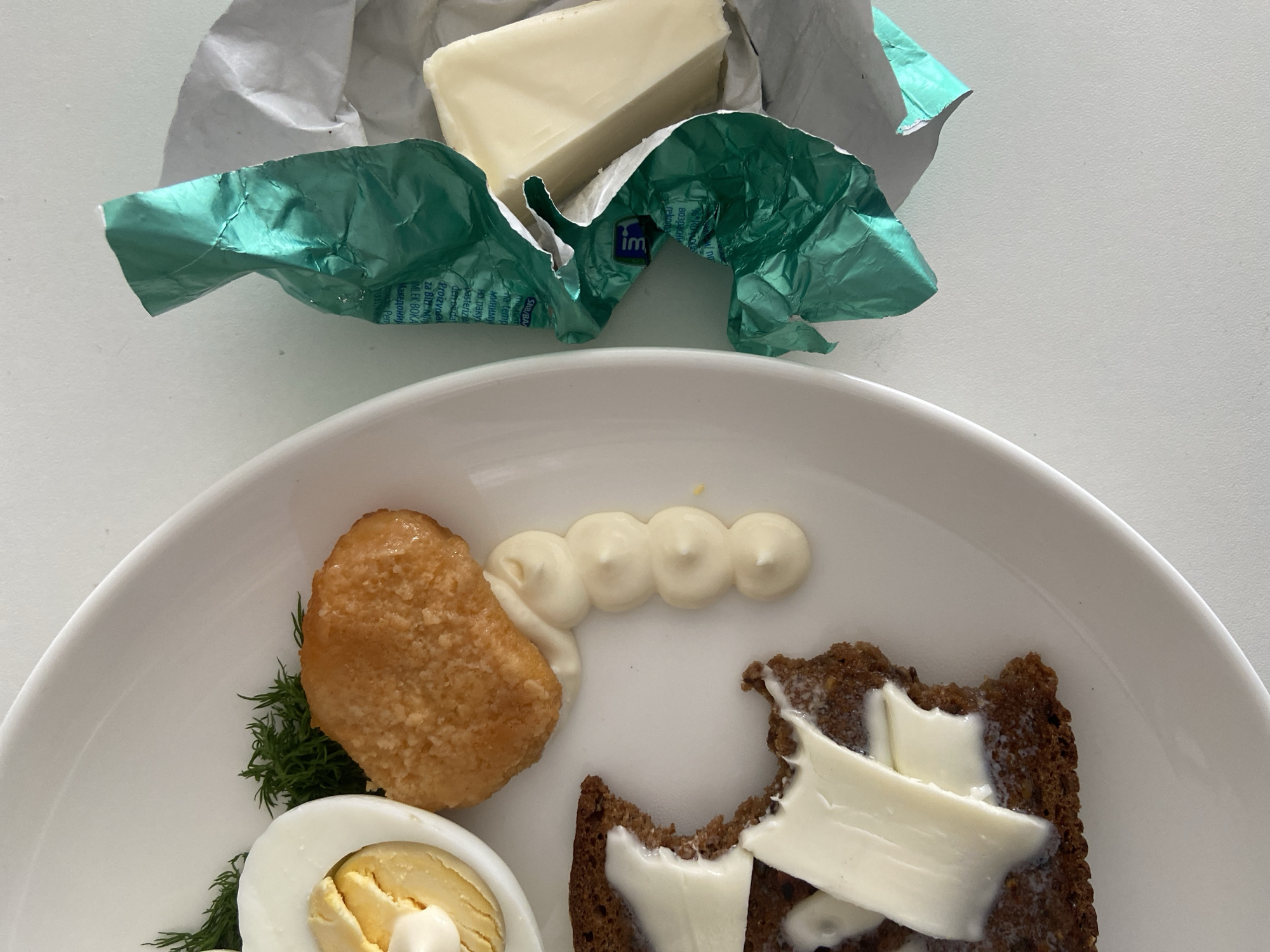 The image depicts a partially eaten meal on a white plate. A piece of dark bread, topped with butter slices, has a bite taken out of it. Next to it is a processed food item, possibly a fish stick or chicken nugget, garnished with a sprig of dill and connected by a line of mayonnaise. A halved boiled egg, also topped with mayonnaise, is present. The wrapper from a butter package, containing the remaining butter, is visible above the plate. The wrapping has the words: "SRB/BA" and "IMLEK BOKA". There is no indication that the image was taken in Budva, Montenegro.