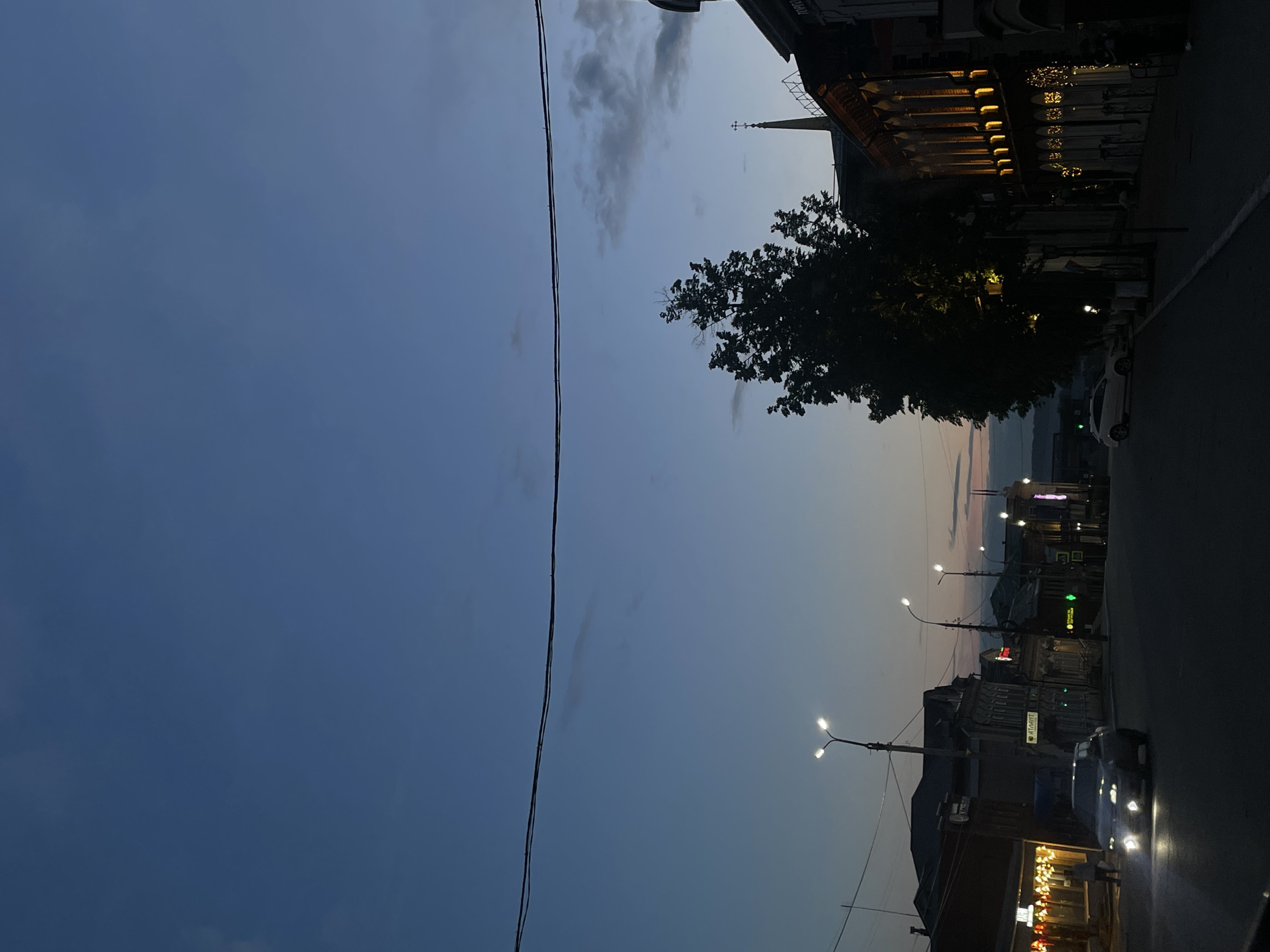 The image captures an evening street scene in Votkinsk, Russia, at dusk or early night. The perspective looks down a street lined with buildings, with a dark blue sky, showing faint clouds and a lighter horizon, dominating the left side of the frame. A thick power line stretches horizontally across the upper middle section of the view.
On the right side of the street, multi-story buildings with varied architecture are visible. One building in the upper right features a facade brightly illuminated by numerous warm lights, suggesting a commercial establishment or a building with prominent architectural details. Further down the street, another building displays a distinct spire or steeple. A large, dark tree with dense foliage stands along the street, partially obscuring the structures behind it.
The street and sidewalks are illuminated by several bright streetlights, casting white-yellow light. Multiple vehicles are present; a white car is parked near the tree, and the headlights of other cars can be seen further down the road. A single pedestrian is faintly visible near a brightly lit storefront in the bottom right corner, walking along the sidewalk.
Visible textual cues include a green cross symbol on a sign, indicating a pharmacy. Another sign features red Cyrillic text, though it is indistinct due to distance and low light. The scene conveys a calm, typical urban evening atmosphere.