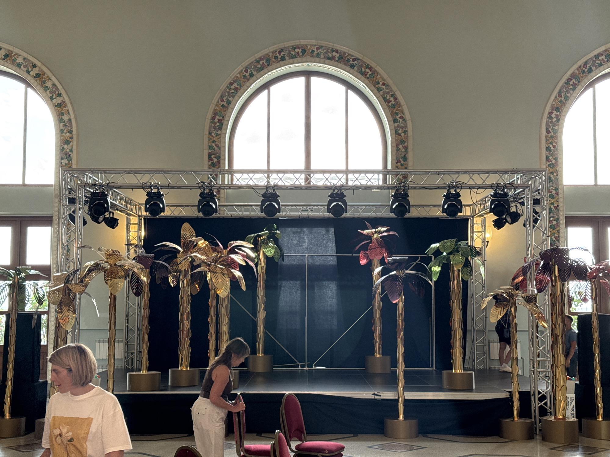 The image depicts the interior of a venue, possibly a convention hall or theater, in Sochi, Russia. The space features a raised stage set up with several large, decorative palm trees. These palm trees are metallic and appear to be golden, with some leaves in shades of green, gold, and red, suggesting a tropical or celebratory theme. A metal truss structure with multiple spotlights is suspended above the stage, indicating preparation for an event.
In the foreground, two individuals are visible. On the left, a woman with short blonde hair and a white t-shirt with a floral design is looking to her left. On the right, another person, partially obscured, is interacting with red upholstered chairs, arranging them near the stage. The chairs themselves are ornate, with dark red velvet upholstery and exposed wooden frames.
The background reveals large arched windows with multiple panes, allowing natural light to enter the space. The windows are framed with decorative tile work featuring floral motifs. The walls are painted a pale green, and the ceiling is partially visible with similar decorative elements around the arches. The overall impression is a venue undergoing setup for an event, with a focus on a tropical aesthetic.