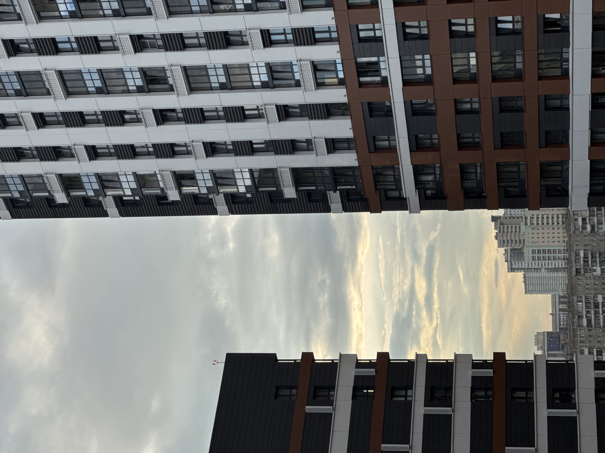The image depicts a city skyline in Novosibirsk, Russia, featuring modern high-rise apartment buildings against a cloudy sky. On the left, a dark grey and white building with horizontal brown accents is prominent, its geometric facade punctuated by numerous windows. To the right, a taller building with a predominantly white exterior is visible, featuring vertical black elements and a repeating pattern of windows with dark frames, some of which are partially open. The lower section of this building is clad in a reddish-brown material. In the background, a cluster of other residential buildings of varying heights and architectural styles are visible, stretching across the horizon. The overall scene is characteristic of urban development in a contemporary Russian city.