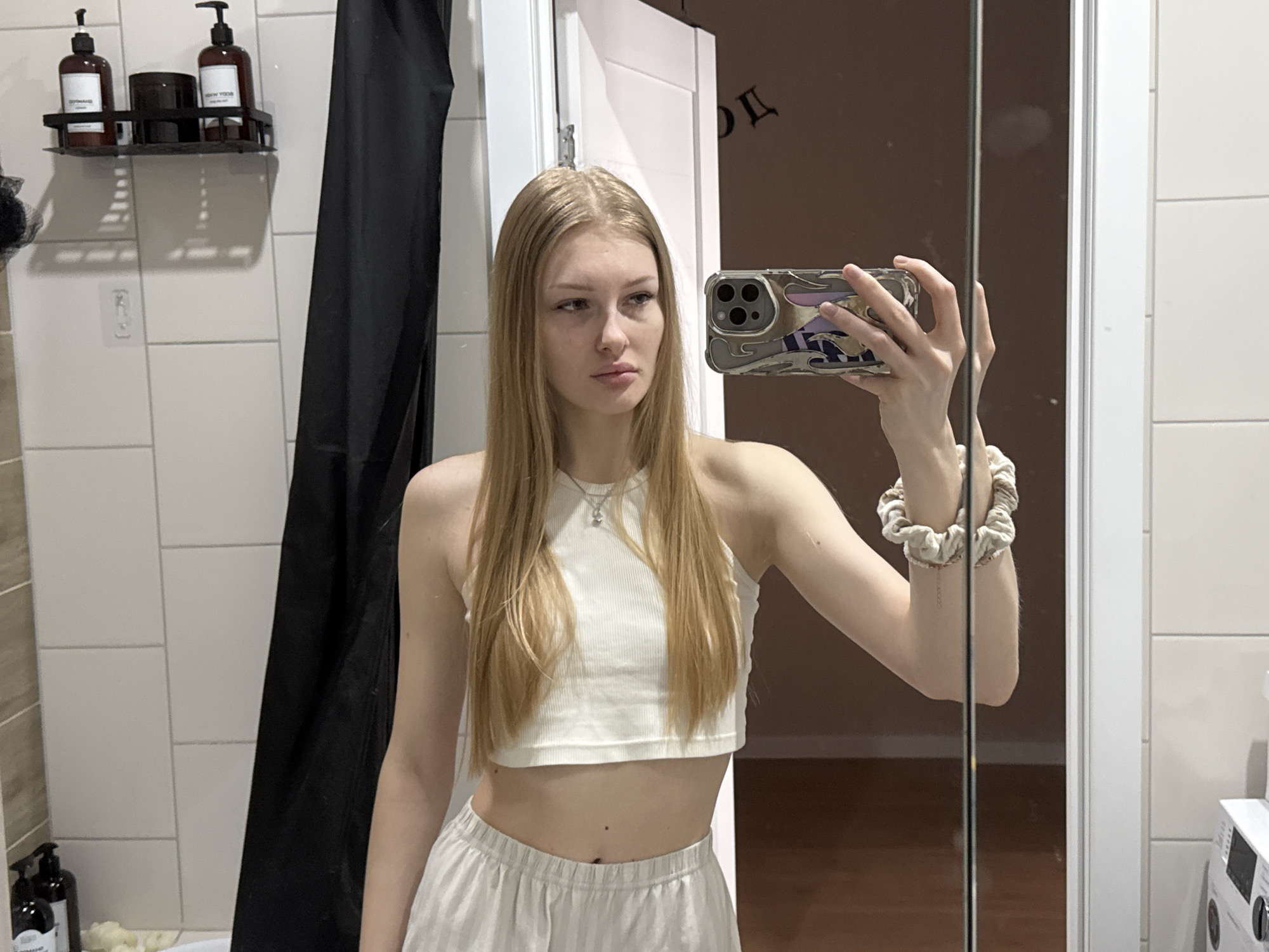 A young woman is taking a mirror selfie in what appears to be a bathroom. She has long, straight blonde hair and fair skin, looking slightly to her left (our right) with a neutral, contemplative expression. She wears a white ribbed crop top, revealing her midriff, and light-colored, possibly striped, wide-leg pants. A silver necklace is visible around her neck, and a light-colored, patterned scrunchie is on her left wrist.
She holds a smartphone with a distinctive, ornate multi-colored case in her right hand to capture the reflection.
The background shows white tiled walls. To the left, a black wall-mounted shelf holds two dark pump bottles and a dark jar. Mirrored text on the bottles faintly indicates "SHAMPOO" and "BODY WASH." A black shower curtain hangs to the right of the shelf. The mirror reflects a room behind the woman with a dark brown wall or door and a wooden floor. On a white door visible in the reflection, faint, mirrored Cyrillic text, possibly "ДГ", can be discerned. A partial view of a white appliance, resembling a washing machine, is also reflected on the far right. The lighting is indoor and ambient. The setting suggests a domestic environment, possibly in Perm, Russia.