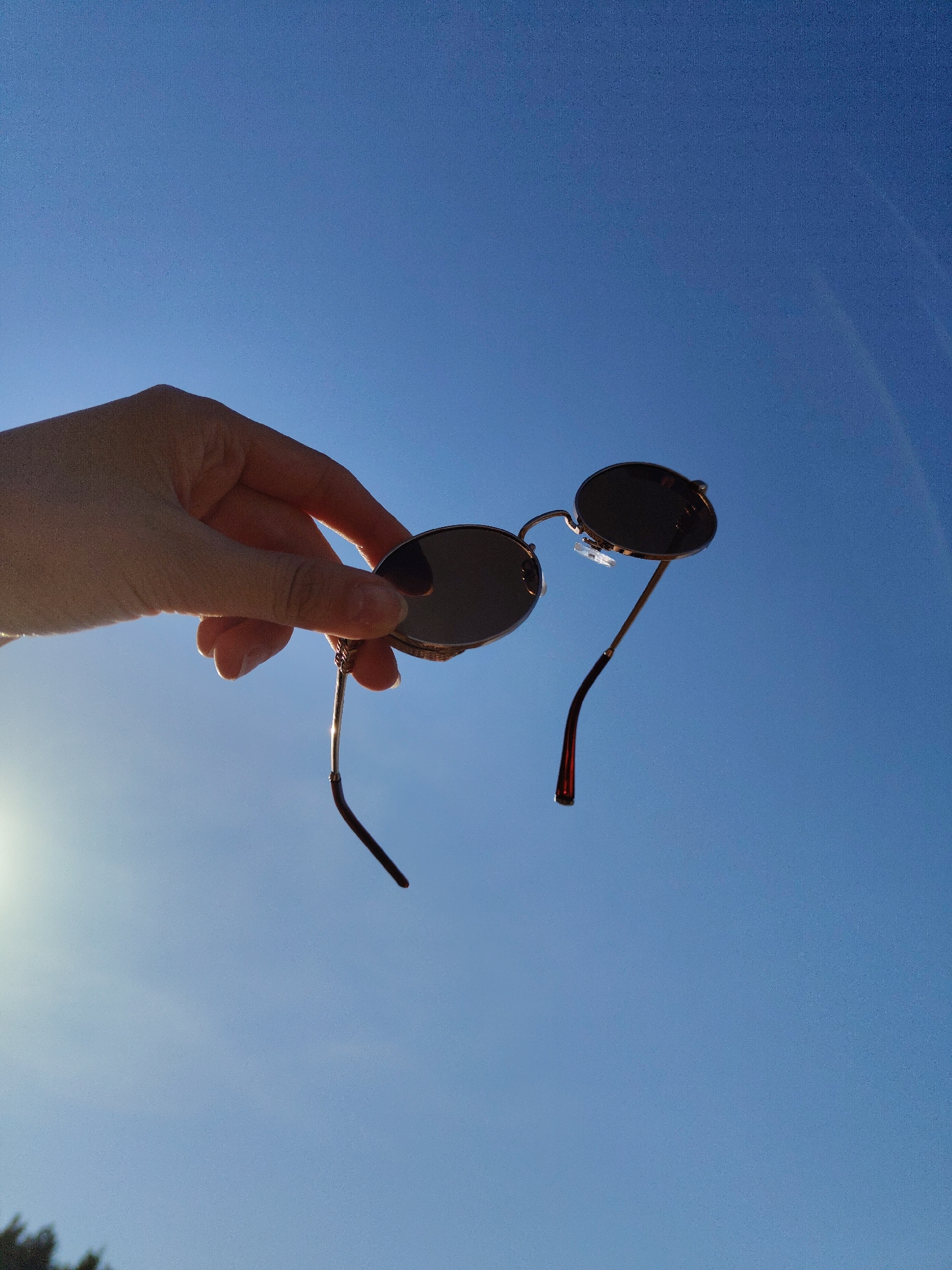 A hand holds up a pair of round sunglasses against a clear blue sky. The sunglasses have dark lenses and thin metal frames. One arm of the sunglasses is dark brown. The sun is faintly visible in the lower left corner of the image. The city and state are not discernable from the image.