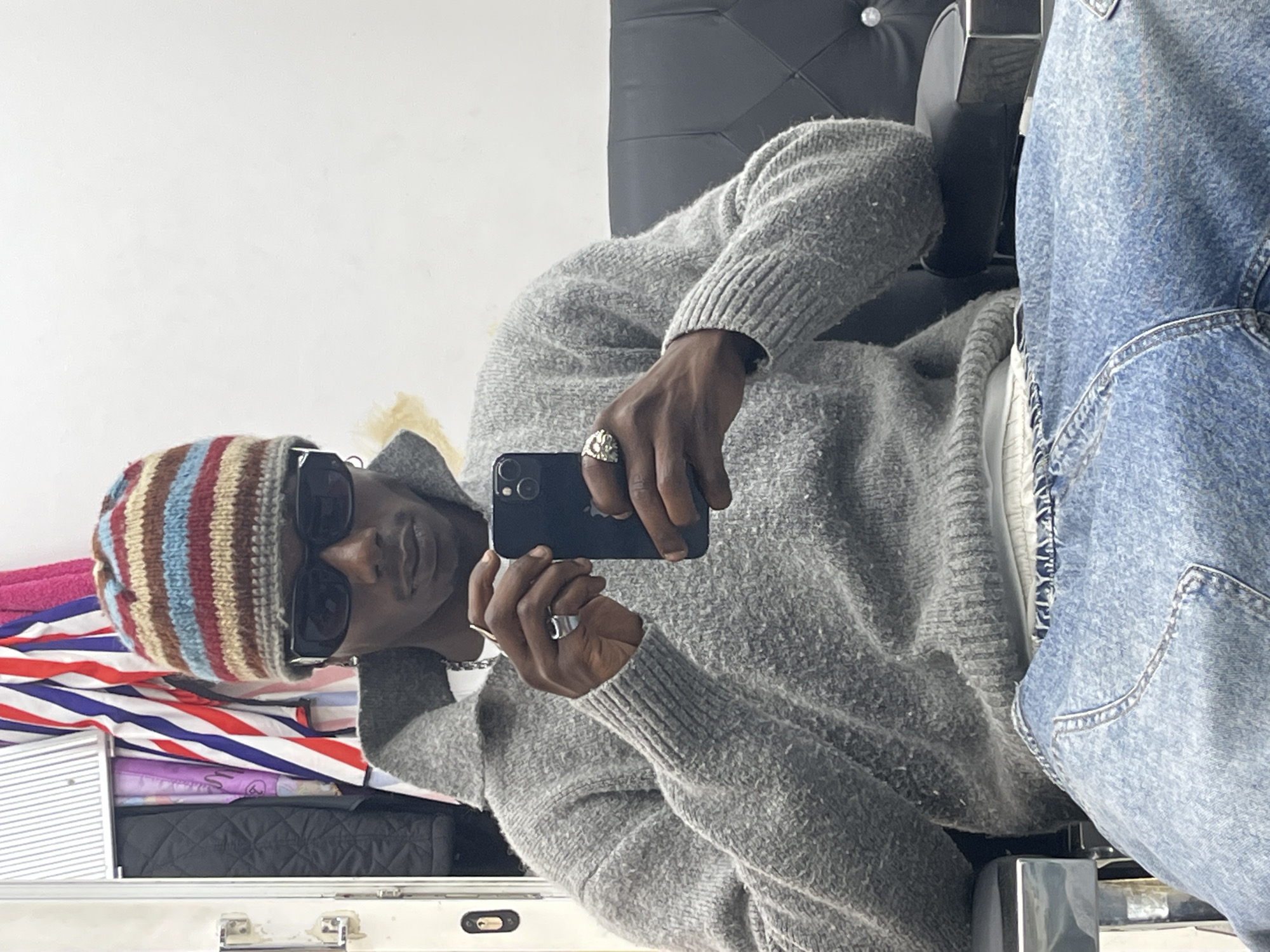 A person with dark skin is sitting in a chair taking a selfie with a black phone. The person is wearing a gray sweater, a striped beanie, sunglasses, rings, and a silver necklace. The chair is black with a silver frame. In the background is a suitcase, and the person is also wearing light-wash denim jeans. The location cannot be confirmed as Barcelona, Spain, based solely on the image.