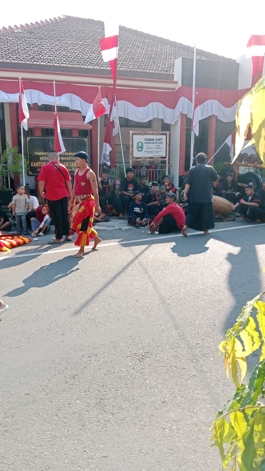 In Magetan, Indonesia, a group of people are gathered outside a building decorated with red and white flags. The building appears to be a government office, indicated by signs that read "PEMERINTAH KABUPATEN MAGETAN" and "KANTOR DINAS TAN...". One man in a red and yellow traditional outfit is walking across the asphalt road, while others are seated on the ground. Some individuals are playing musical instruments, including a drum. The sun is shining, casting long shadows on the road. The scene suggests a cultural event or celebration taking place.