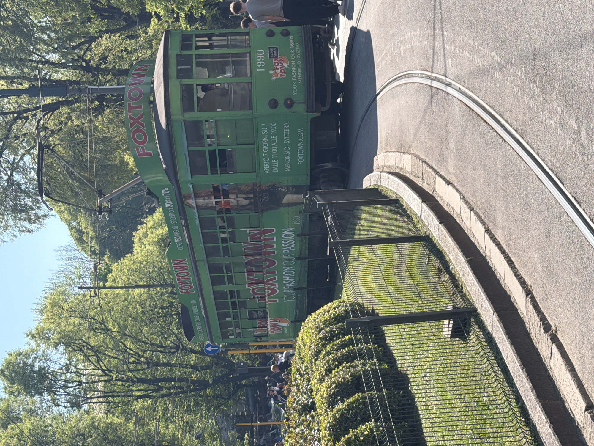 A green and red streetcar is seen on a tram track curving to the right. The streetcar has advertisements for FOXTOWN, a factory outlet store in Mendrisio, Switzerland. It states it is "OPEN 7 DAYS" with the hours of 11 AM to 7 PM. The streetcar is labeled "1990," and the website foxtown.com is shown.
People are standing on the sidewalk next to the streetcar. Trees and foliage are visible in the background. A black metal fence separates the streetcar tracks from a grassy area. A street sign is also present, indicated by a blue arrow. The location is likely in Milan, Italy.