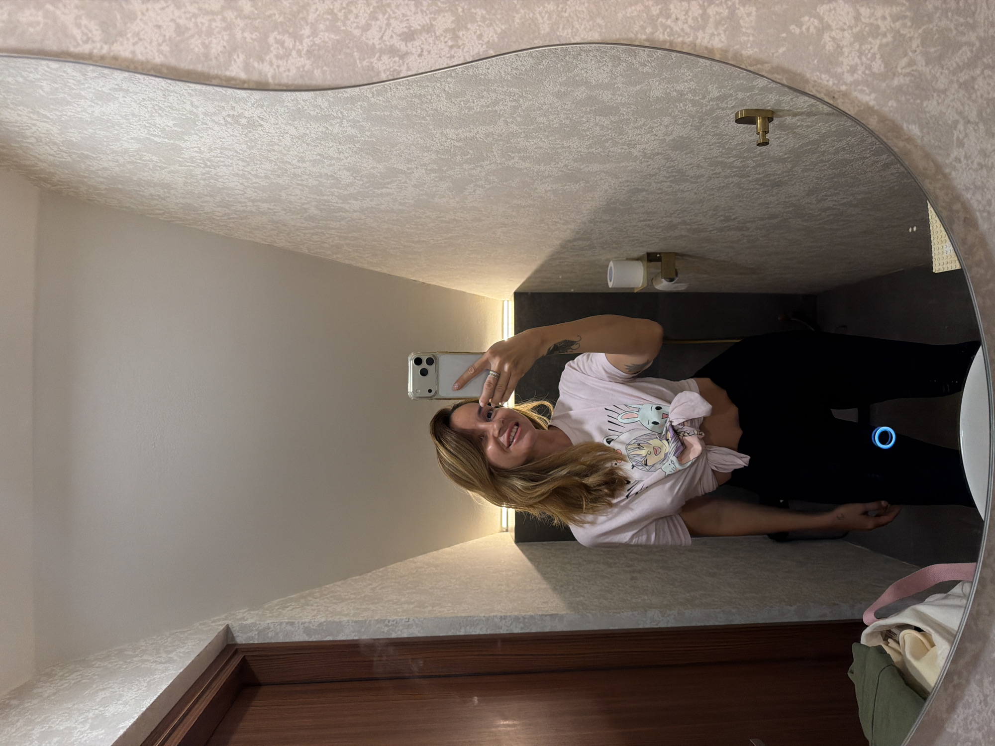 A woman is taking a mirror selfie in a bathroom. She is wearing a pink t-shirt with a cartoon graphic and black pants. The bathroom has textured wallpaper, a wooden door, and a large, irregularly shaped mirror. There is a roll of toilet paper on a holder to her right. On the left side of the mirror, a pile of clothes is visible in the foreground. The lighting in the bathroom appears to be dim, with a bright light source illuminating the wall behind the woman. The scene is likely indoors, possibly in a hotel room or a private residence, and the mention of Kuta Selatan, Indonesia, provides a geographical context for the location.