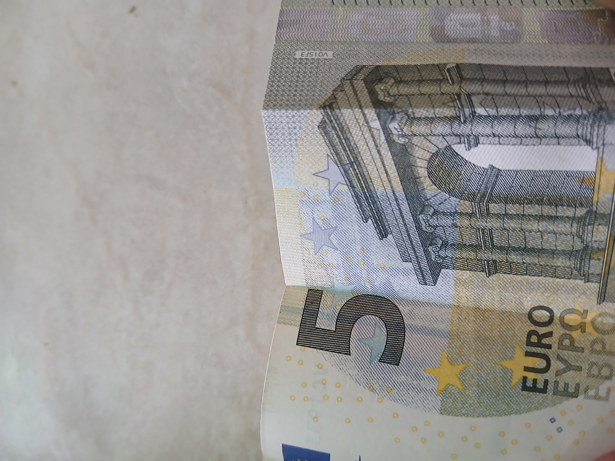 The image shows a close-up of a five Euro banknote. The note displays the numeral "5" and the word "EURO" printed on it. A depiction of architectural elements, possibly an archway or doorway, is visible on the bill. The background appears to be a light-colored surface. There is no indication of the location being in Juan Griego, Venezuela.