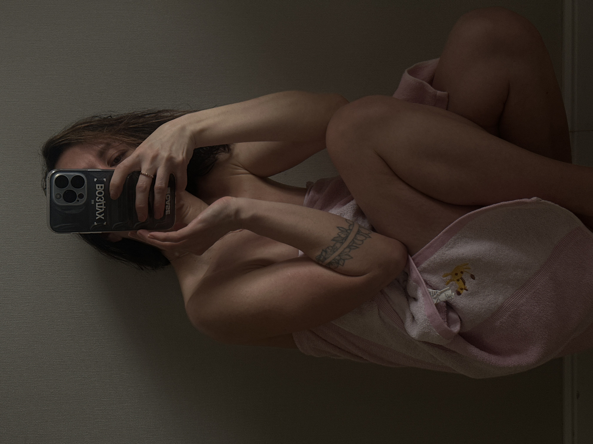 A person is depicted in a sideways, full-body mirror selfie against a plain, light grey wall, likely indoors. The individual, who appears to be female, is partially nude, wrapped in a light pink towel that features an embroidered yellow giraffe design. The person's face is obscured by a smartphone, held up to capture the reflection. The smartphone, an iPhone model with multiple cameras, is encased in a clear case. Visible text on the phone case, appearing reversed in the mirror, includes "BO3TЛXX" and "MICASE". A ring is worn on the right hand, and a tattoo, resembling a floral or vine pattern, is visible on the left arm. The person is posed in a reclined or seated position, with legs bent and tucked. The lighting is dim, creating a soft, intimate atmosphere.