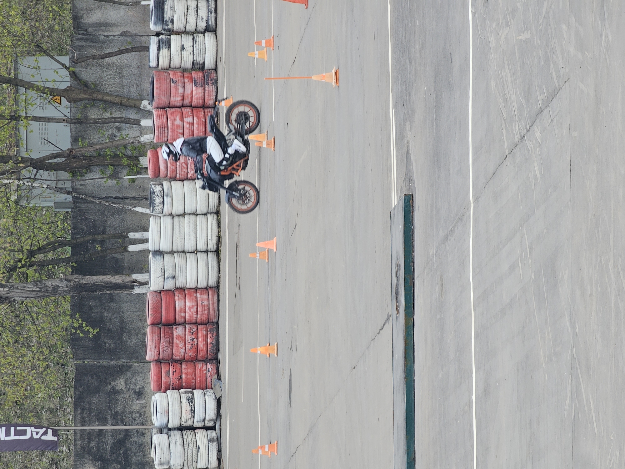 An overhead, vertically-oriented shot captures a motorcyclist on a paved asphalt track, likely a training ground or a closed course. The rider, dressed in a white helmet, black jacket, and white pants, is positioned on a black and orange motorcycle, seemingly in motion or preparing for a maneuver.
To the left of the track, a protective barrier is formed by neatly stacked used tires, alternating in color between white/grey and red. Further to the left, green trees with visible branches and a textured concrete wall can be seen. A utility box with a yellow warning sign is partially visible among the trees.
Scattered across the asphalt surface are several bright orange traffic cones, indicating a designated path or obstacles for training. White lines are painted on the asphalt, delineating lanes or sections of the course. The track also shows signs of wear, including cracks and patches.
The scene is set outdoors during daylight hours, under bright, clear conditions, suggesting fair weather. In the bottom left corner (when oriented correctly), a black banner or flag is visible with the word "TACTIC" printed in white. The overall impression is of a motorcycle skills training or practice session taking place in an urban or semi-urban environment.