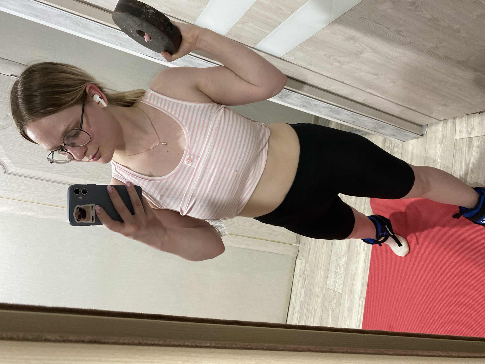 A woman is seen in a mirror selfie. She has blonde hair, glasses, and is wearing a striped pink and white tank top and black biker shorts. She is holding a weight in her raised right hand and a cell phone in her left hand. The phone has a blue case with a cartoon image on it. She is wearing an AirPod in her ear. On her ankles, she is wearing blue ankle weights. She is standing on a red yoga mat. The background includes a light wooden door or cabinet. The photo was likely taken indoors. The location is Chaikovsky, Russia.