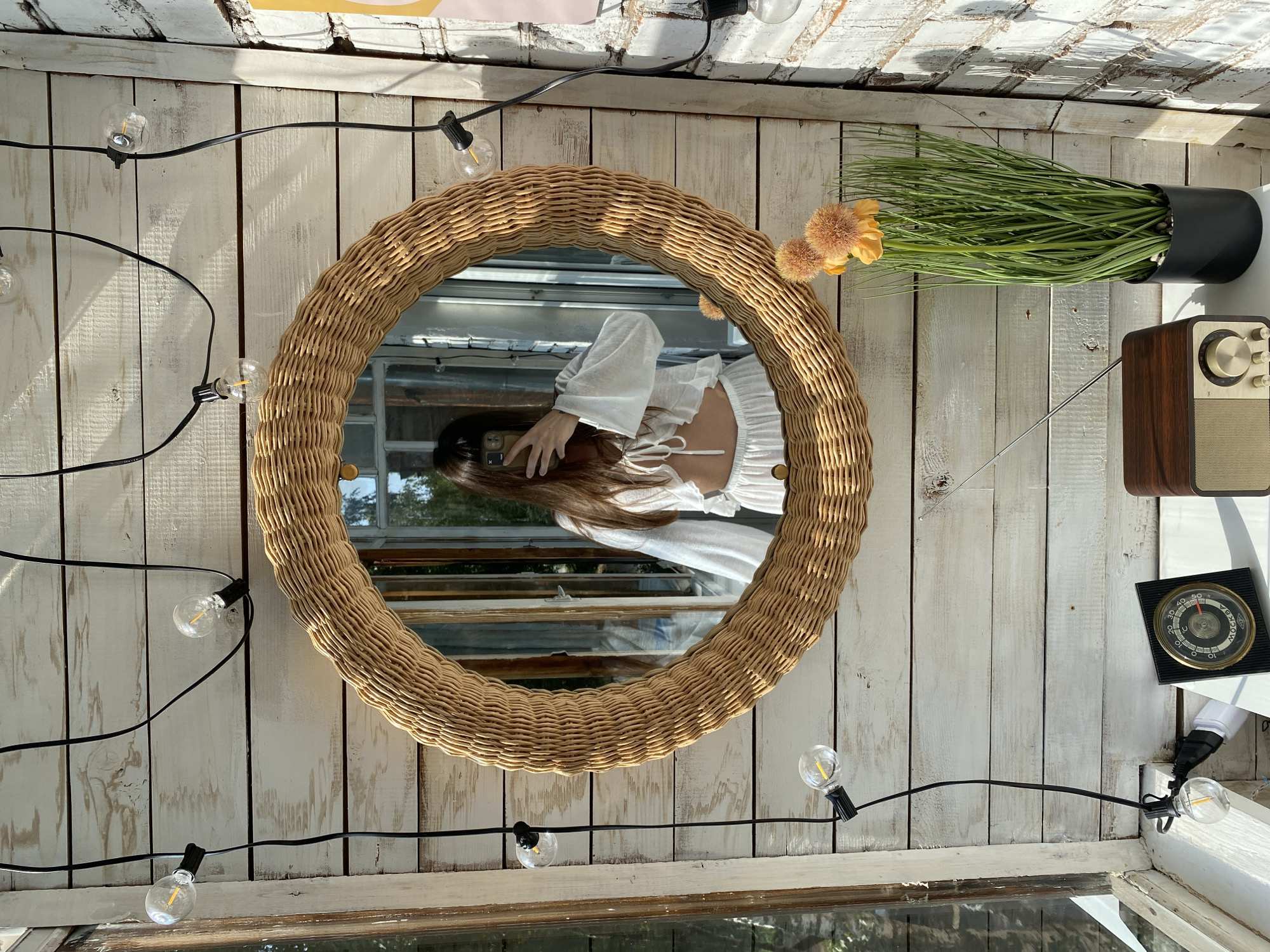 A woman with dark hair is taking a selfie in a round wicker mirror. She is wearing a white two-piece outfit. The mirror is hanging on a wall made of white-painted wooden planks. String lights with clear bulbs are draped around the mirror and along the wall. To the left of the mirror is a window with sunlight streaming in. Below the mirror, on a white surface, are a vintage-looking radio and a black clock with a circular face. To the right of the radio is a potted artificial plant with long green grass-like leaves and two orange pom-pom flowers. The scene appears to be in a sunroom or a covered outdoor space. The location is identified as Votkinsk, Russia.