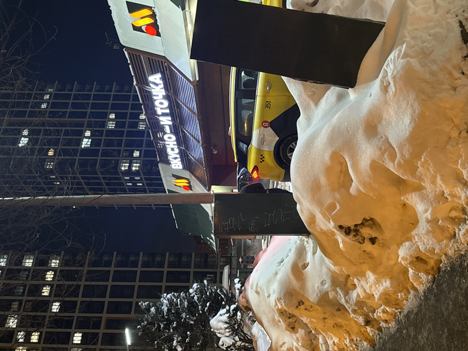 A sideways shot shows a Vkusno - i tochka restaurant at night. A yellow taxi with the number 351 is parked beside it. In the foreground, a large pile of snow dominates the frame. A tall building with illuminated windows is visible in the background. A tree covered in snow is in front of the building. The sky is dark.