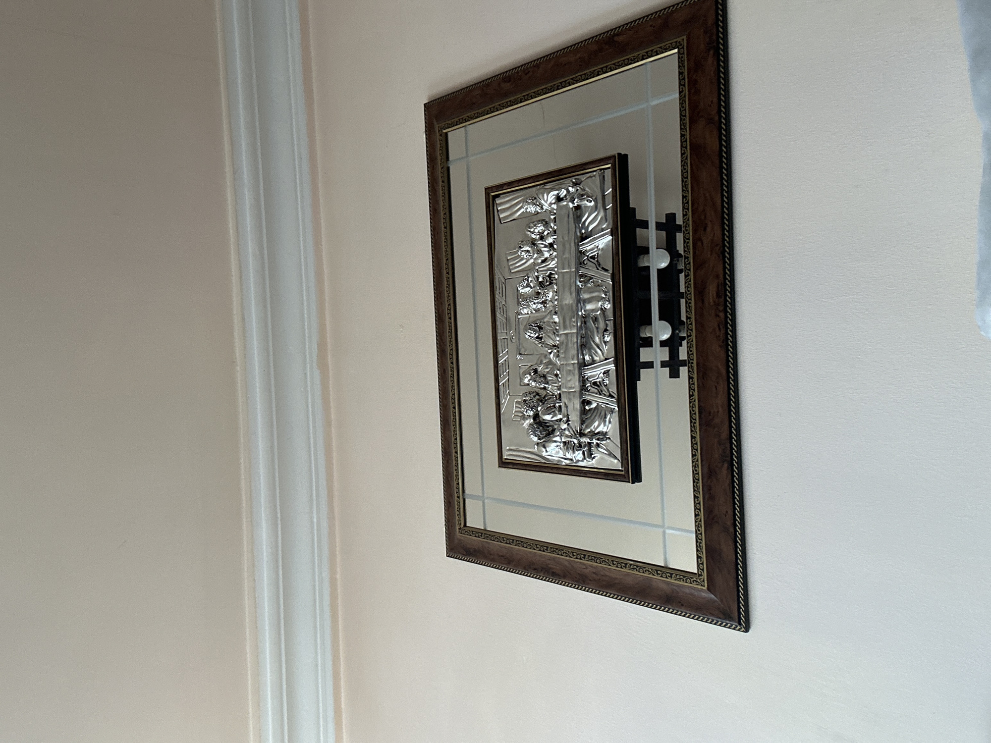 The image shows a framed mirror with a smaller framed image of a relief sculpture hanging on a light-colored wall. The wall is painted a pale beige or off-white color. To the left of the frame is a white architectural moulding, potentially a door or window frame. The large frame is dark brown and has an ornate design. The hanging mechanism is visible behind the right side of the framed picture.