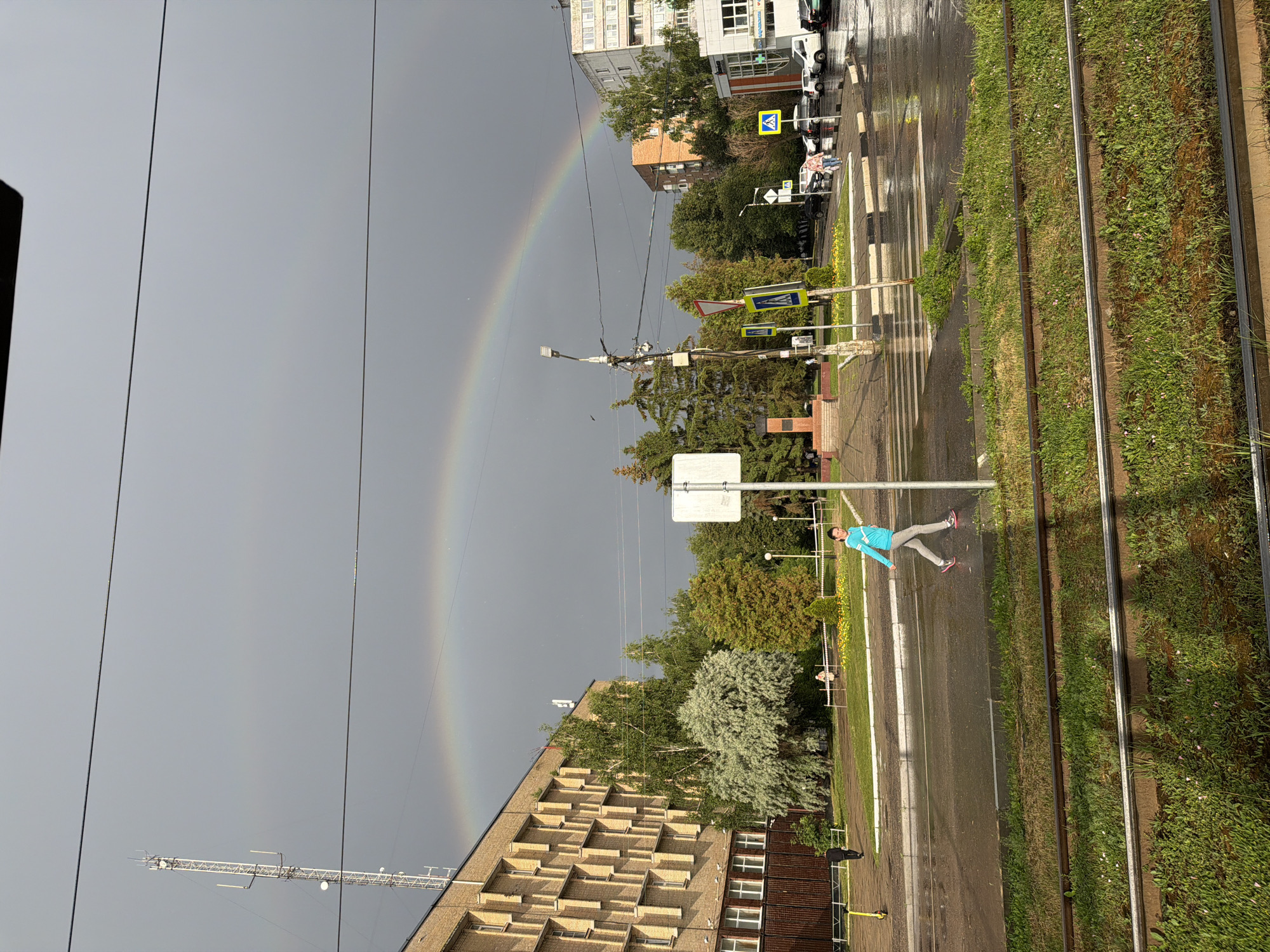 The image depicts a post-rain scene in Kolomna, Russia, with a prominent rainbow arcing across a grey sky. In the foreground, a person in a blue jacket and grey pants is captured mid-stride, seemingly enjoying the wet pavement and the colorful sky. The setting is an urban environment with tram tracks running parallel to the road, bordered by lush green grass. Several buildings, including a multi-story residential or office building with a distinct brick facade, line the street. Street signs, including a yellow pedestrian crossing sign, are visible. The wet road surface reflects the subdued light. The time of day appears to be late afternoon or early evening, judging by the light and the presence of a rainbow, which often appears after rain when the sun is low in the sky. The overall mood is serene, with a touch of wonder from the natural phenomenon. No legible text is discernible in the image.