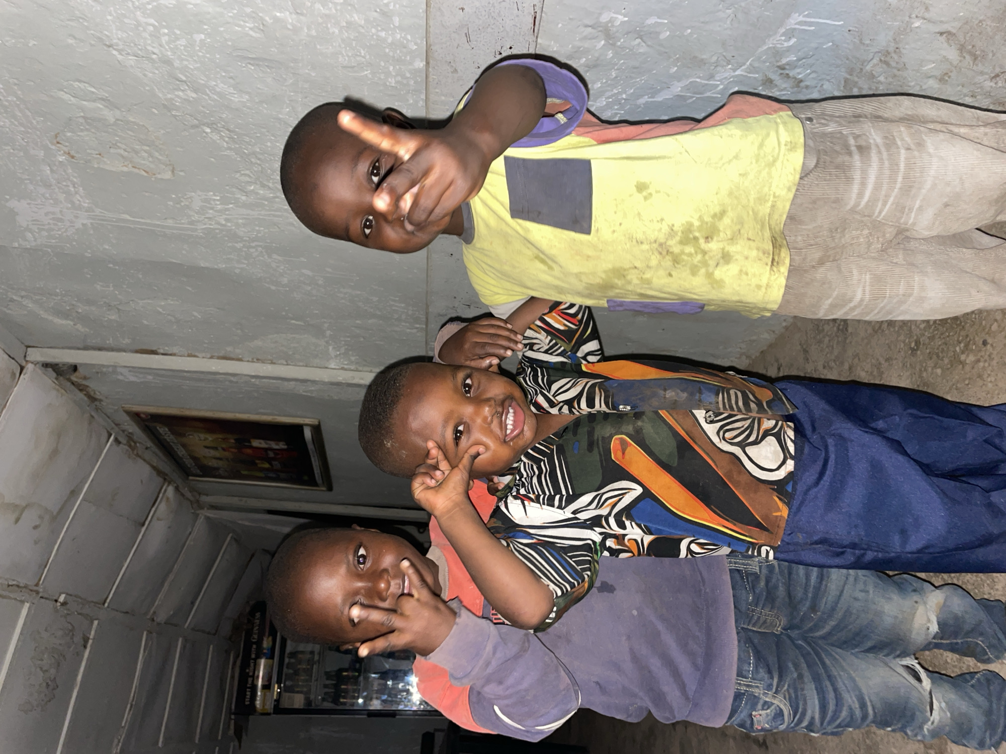Three young children are posing for a photo. They are indoors, possibly in a bar or shop in Jos, Nigeria. The child at the top is wearing a yellow and purple shirt and khaki pants and making a peace sign. The middle child is wearing a patterned shirt and jeans and also making a peace sign. The bottom child is wearing a purple and red shirt and jeans and making a peace sign as well. Behind them, on the left, there is a framed picture on the wall. A cooler is behind the bottom child. The walls are gray and textured.