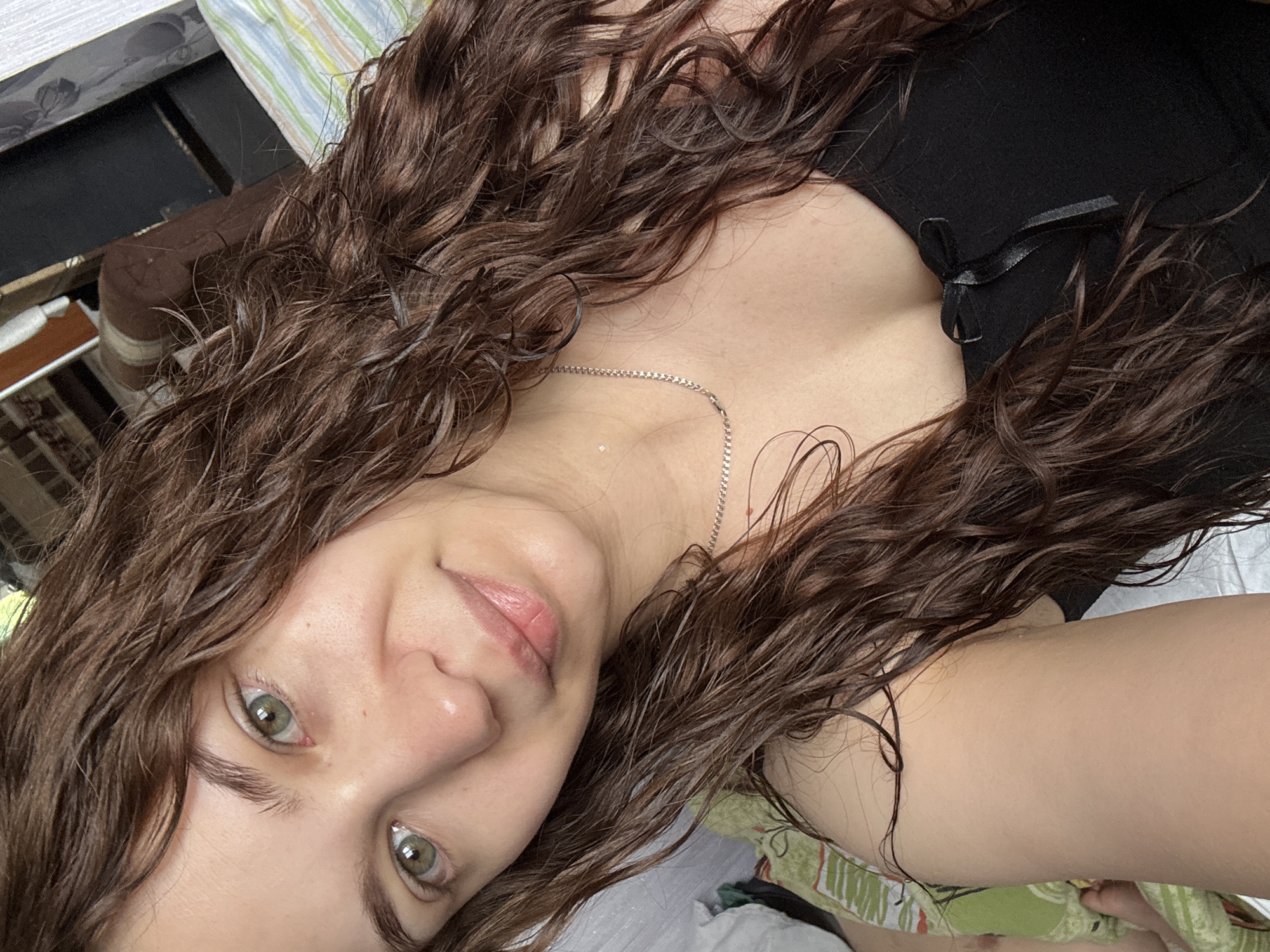 A selfie shows a woman with long, wet, curly brown hair. She has fair skin and green eyes. She is wearing a black top with a small bow at the neckline and a silver chain necklace. The background includes a bed with striped bedding, and a dark piece of furniture. There is no indication of the location in the city of Nizhnyaya Gar', Russia.