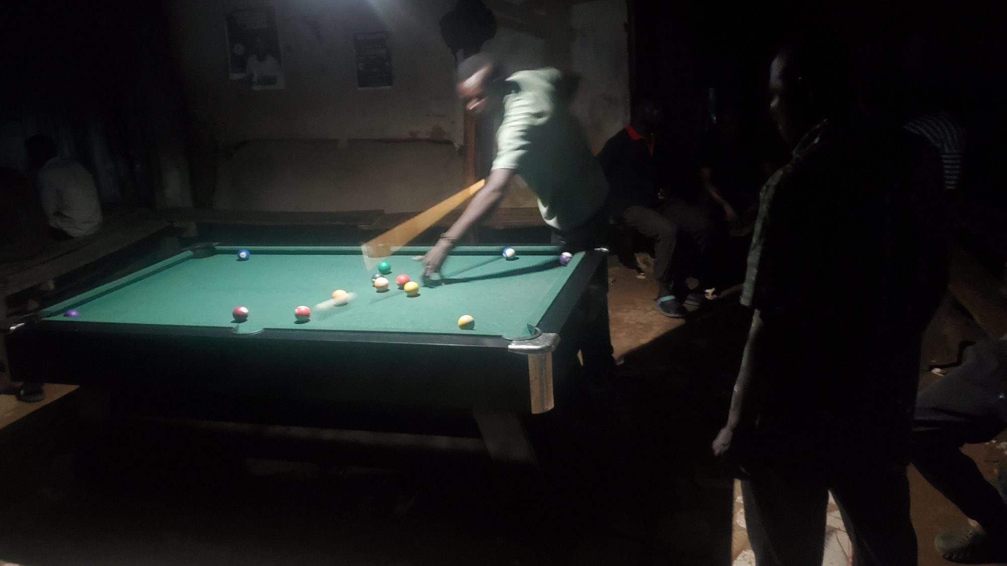 The image depicts a dimly lit room, likely a bar or recreational space, in Jalingo, Nigeria. A group of men are gathered around a green pool table. One man in a green shirt is actively playing pool, taking a shot with a cue stick. Several billiard balls are scattered across the table, including a blue ball, several solid colored balls (red, yellow, orange), and what appears to be a striped ball. Other individuals are seated and standing around the table, observing the game. The environment is rustic, with bare walls and simple furnishings, suggesting a casual, local establishment. The lighting is low, creating shadows and highlighting the pool table and the players.