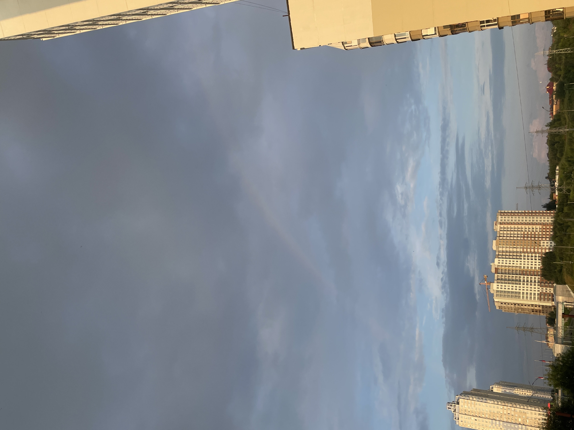 The image captures a sky filled with dramatic clouds, some dark and gray, others illuminated by sunlight, creating a contrast of light and shadow. A faint rainbow is visible in the upper-middle portion of the sky. In the foreground, on the right side, a tall, modern apartment building with a beige and white facade dominates the view. The building features numerous balconies. On the left side of the image, two other tall apartment buildings are visible. One, on the far left, is a slender, light-colored building with many windows. The other, closer to the center, is a wider, light-brown building with a construction crane at its top. Below the buildings, a line of trees forms a green border. Power lines and transmission towers are scattered across the landscape. The scene appears to be an urban or suburban environment, likely in Perm, Russia, given the architectural style and infrastructure. The lighting suggests it is either early morning or late afternoon, with the sun casting a warm glow on the side of the building. No people are visible in the image.