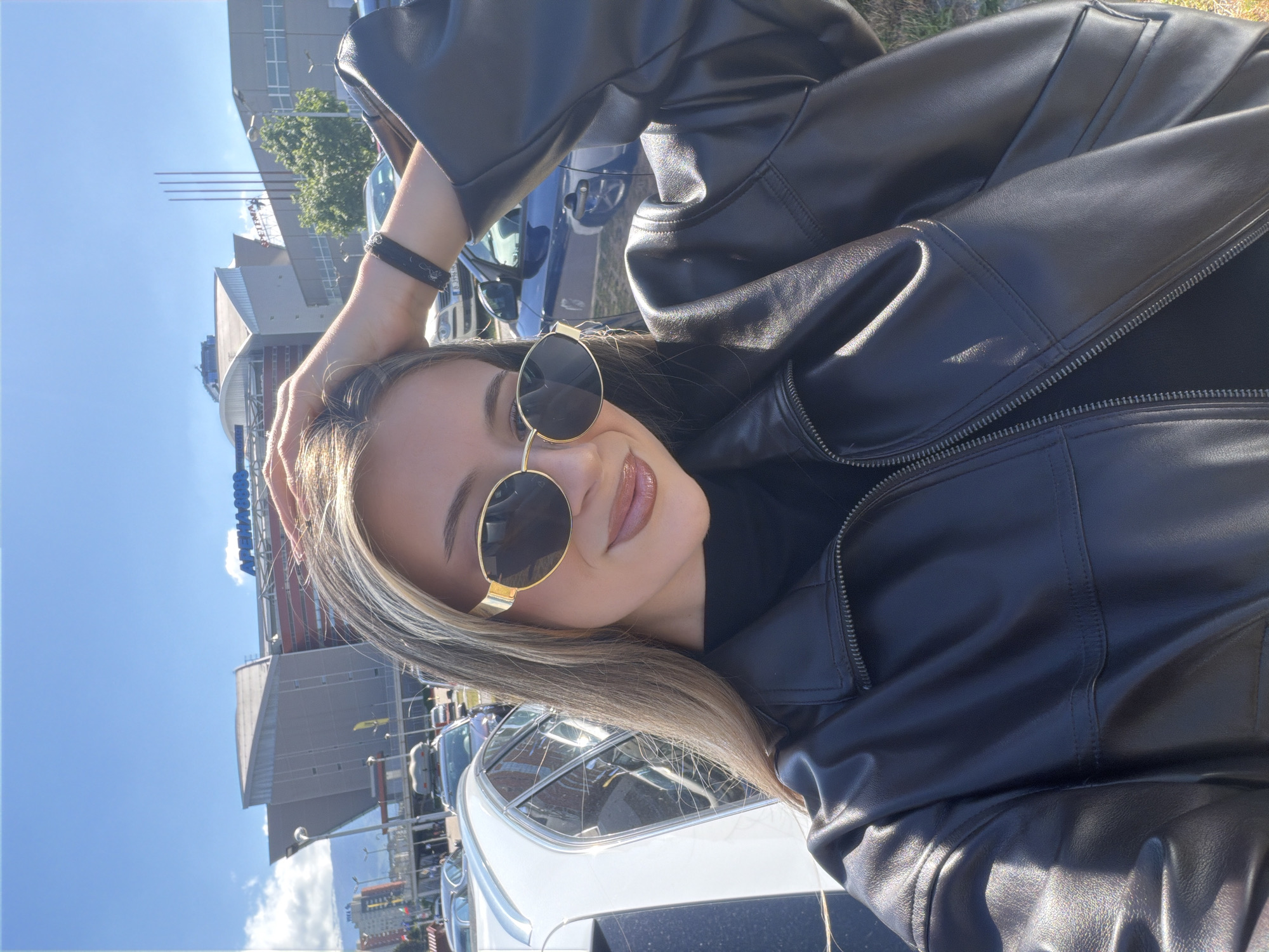 The image shows a woman wearing a black leather jacket and sunglasses, posing with her hand on her head. In the background are vehicles and a large building. The building is identified as "Arena Armeec," placing the location in Sofia, Bulgaria. The weather appears sunny with a blue sky visible.