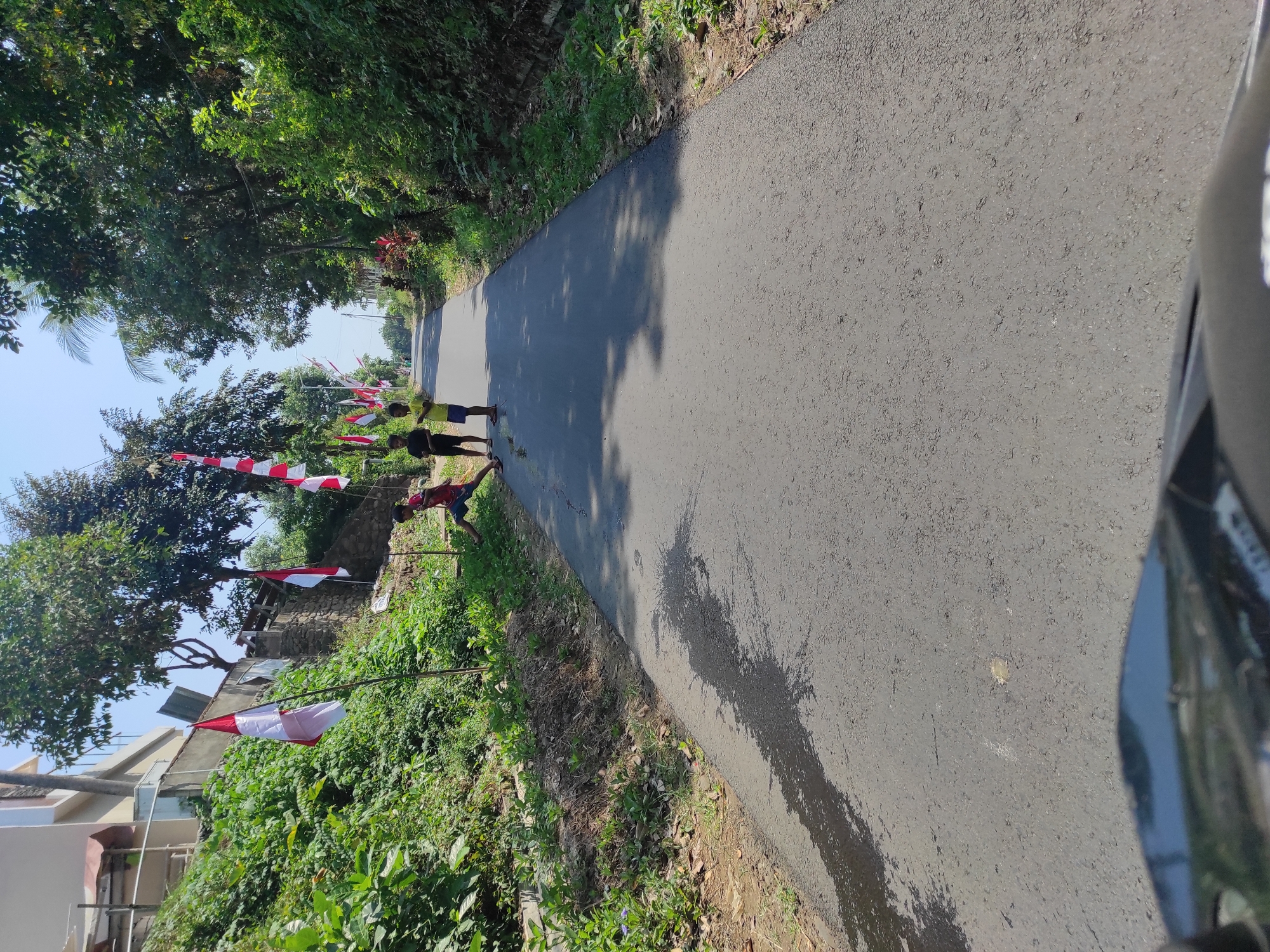 In an outdoor, daytime scene on an asphalt road in an unknown city, several children are present. Three children are visible on the left side of the road. One child, wearing a red shirt and blue shorts, is leaning against a wall. Two other children, one in a black shirt and the other in a yellow shirt with blue shorts, are standing near the roadside. Indonesian flags, red and white, are planted along the side of the road. The road is bordered by lush green vegetation on both sides, with trees and bushes creating a natural environment. The sky above is clear and blue. The foreground of the image shows the edge of what appears to be a vehicle, possibly a motorcycle, suggesting the perspective is from someone riding.