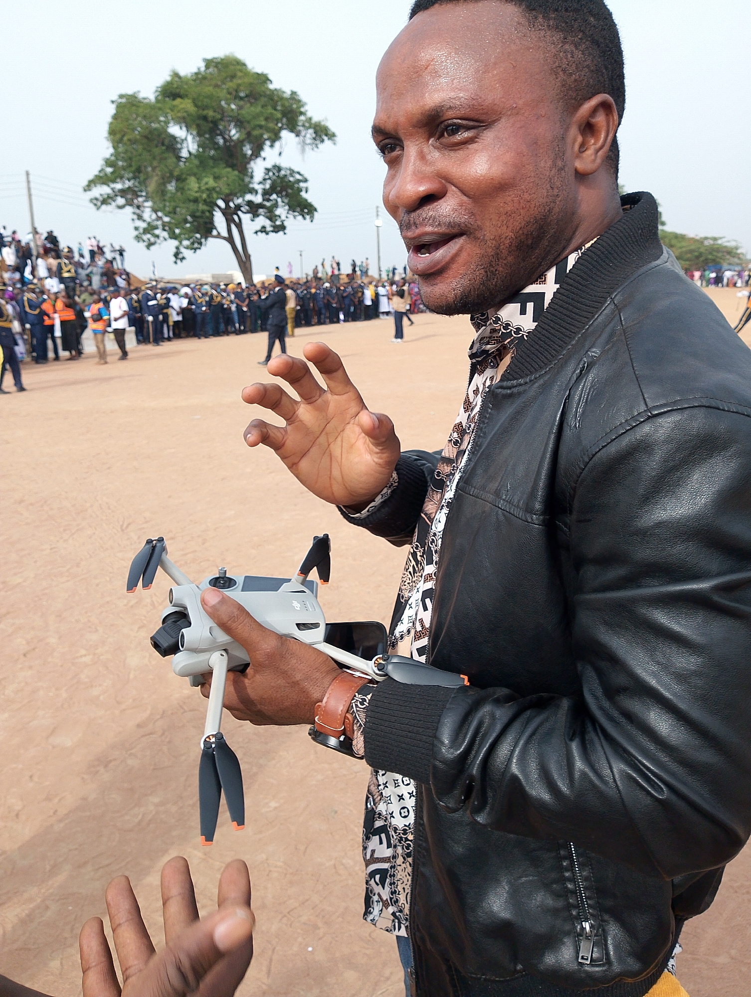 A man with dark skin, wearing a black leather bomber jacket over a patterned shirt, is captured in a close-up, looking to his right with an animated, open-mouthed expression, as if speaking or explaining. He holds a light grey DJI Mini 3 Pro drone with folded propellers in his left hand, along with an attached smartphone that serves as a controller display. His right hand is raised and open, gesturing, while another person's hand is partially visible in the lower left, extended towards the drone, possibly in interaction. The man's shirt features a repeating pattern of dark and light geometric shapes, including what resembles stylized "X" and "O" symbols.
The scene unfolds in a large, open, unpaved area with light brown, sandy ground, typical of an outdoor public gathering. In the background, a substantial crowd of people is gathered, with many individuals wearing dark uniforms, suggesting an official event or ceremony. A prominent, large tree with dense green foliage stands in the mid-background to the left. The bright, slightly hazy sky indicates daytime, likely late morning or early afternoon. The man appears engaged and enthusiastic, seemingly demonstrating or presenting the drone to others. The event is taking place in Kubwa, Nigeria.