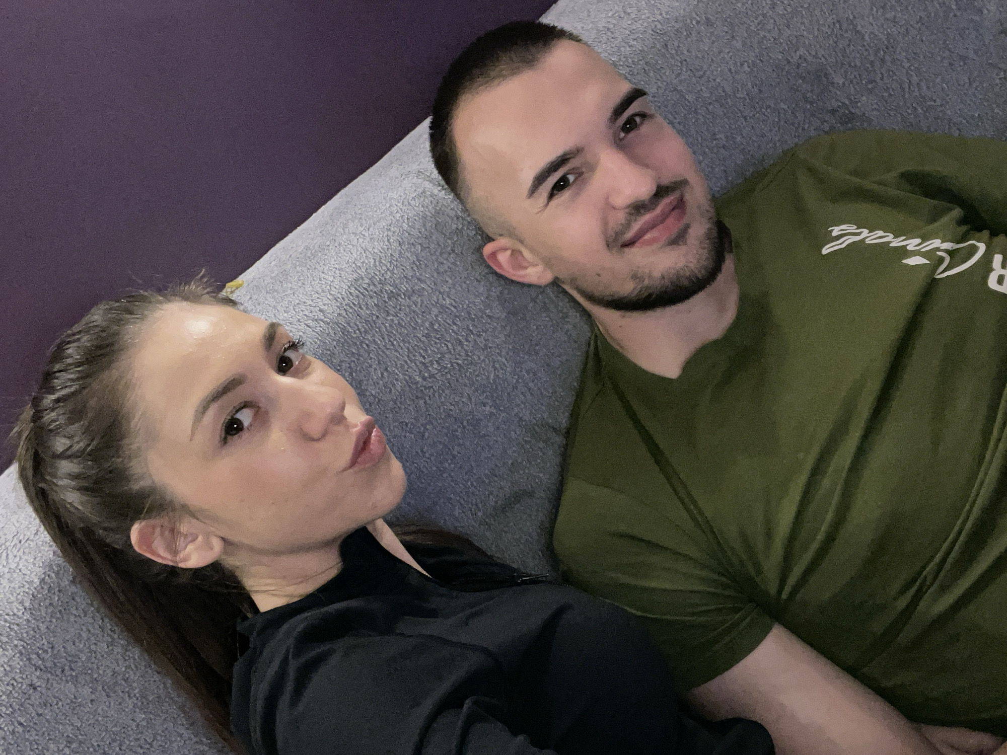 A photo taken at an indoor location, a woman and a man are lying on a gray couch. The woman, on the left, has dark hair pulled back in a ponytail and wears a black zippered jacket. She is looking towards the camera with a pout. The man, to the right, has short dark hair and a beard and is wearing an olive-green t-shirt. The location is not identifiable as being in Sofia, Bulgaria.