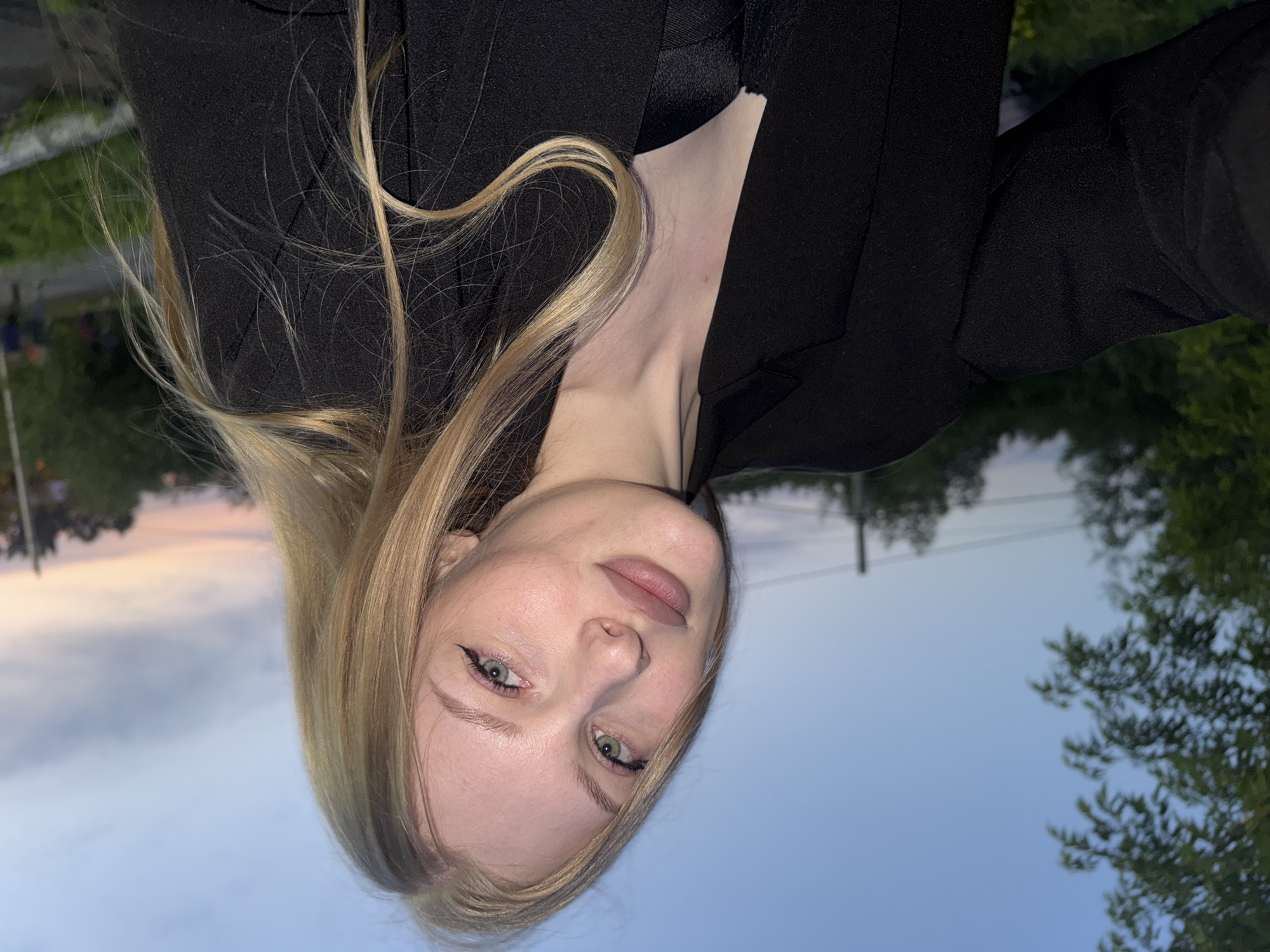 A young Caucasian female with blonde hair and green eyes is pictured from the shoulders up, looking towards the camera. She is wearing a black blazer over a black garment. The background features a dusky sky with a hint of sunset colors, trees, and a few indistinct figures in the distance. The image appears to have been taken outdoors in an urban or semi-urban setting. The location is specified as Perm, Russia.