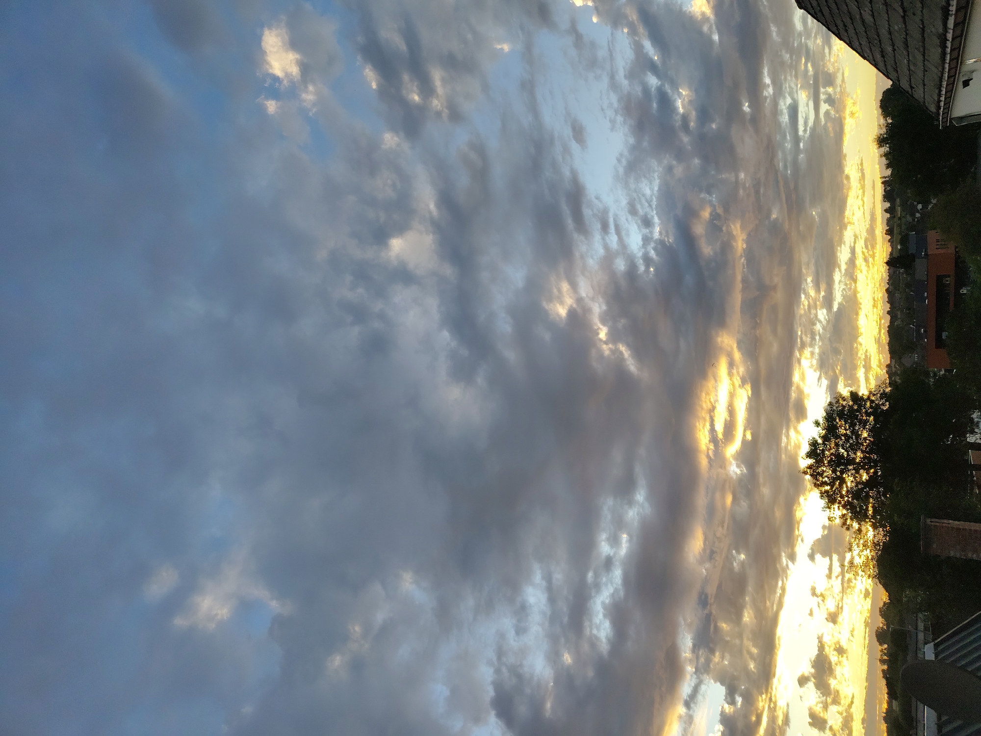 The image depicts a cloudy sky over the city of Nederbrakel, Belgium, likely during sunset or sunrise. The upper portion of the sky is dominated by large, grey clouds, interspersed with patches of lighter blue sky. Towards the horizon, the clouds are illuminated by warm, golden light, suggesting the sun is low in the sky.
In the foreground, the roofs of buildings and a few trees are visible. The roof on the far right appears to be tiled, with a dormer window. To the left, other building structures and greenery are present. A large, silhouetted tree stands prominently in the mid-ground, its branches etched against the bright sky. The overall scene captures a natural, atmospheric moment in the Belgian landscape. There are no people or animals visible in the image, and no discernible actions or interactions are taking place. The environment is that of a developed area, likely residential or a small town, given the presence of buildings and infrastructure.