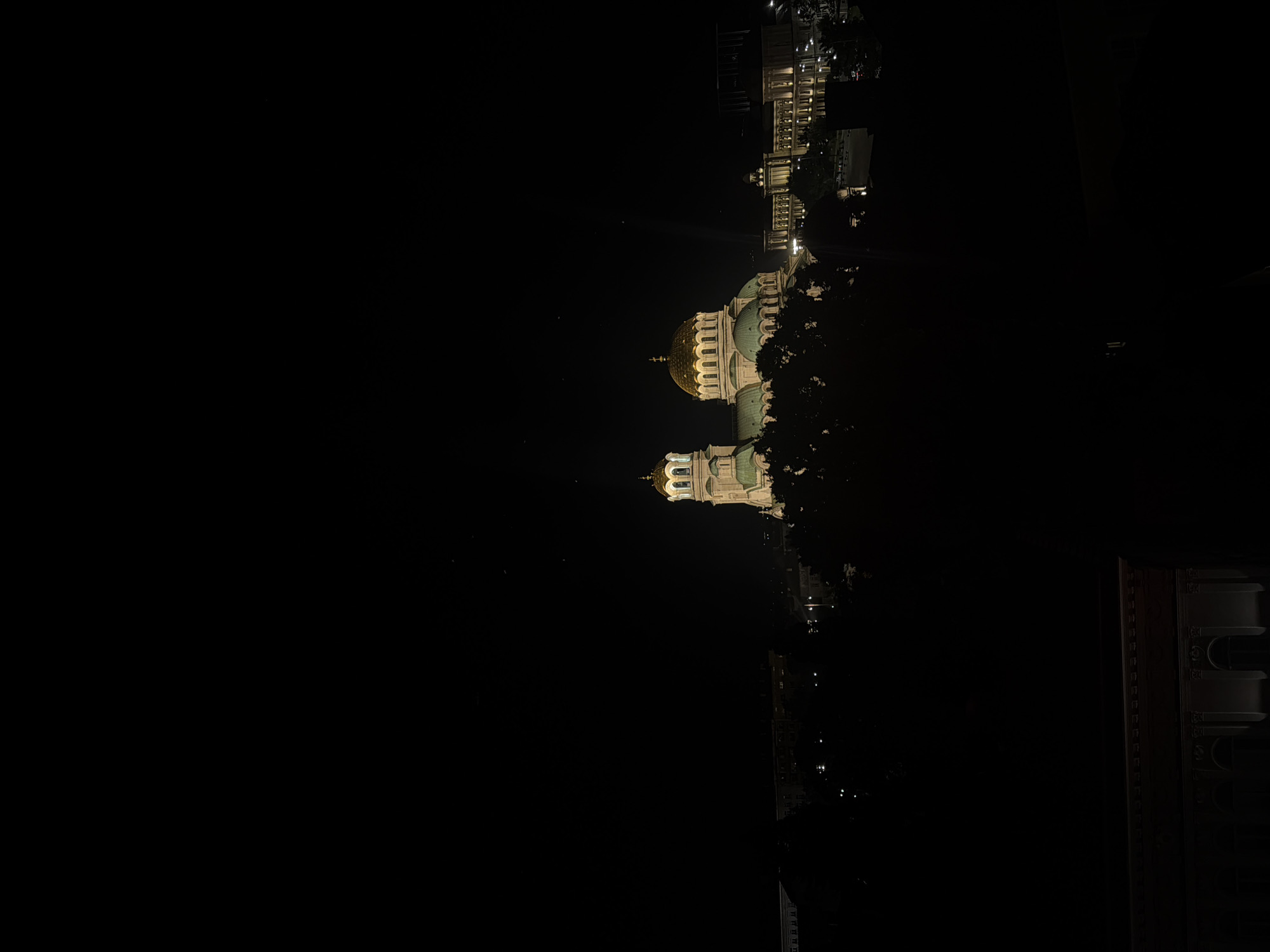 The image is a nighttime aerial view of the Alexander Nevsky Cathedral in Sofia, Bulgaria. The cathedral's golden domes and illuminated facade are the most prominent features against the dark sky. Surrounding the cathedral are other buildings, also lit, and silhouetted trees. The overall scene is dark, with the artificial lights of the city providing the only illumination. No people are visible in the image.