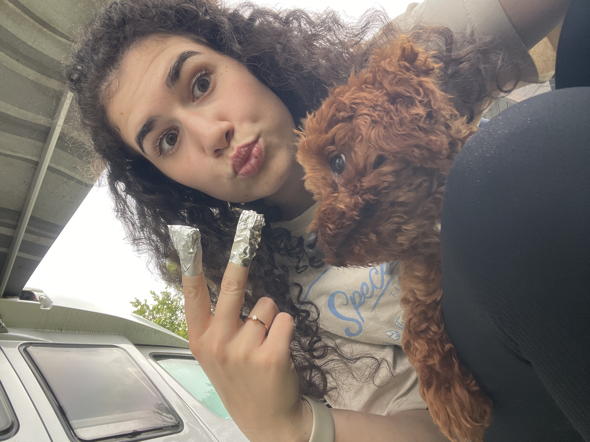 A female with dark, curly hair and light complexion is depicted in a selfie, taken from a low angle. She exhibits a playful expression with wide eyes and puckered lips, directed at the camera. Her left hand is raised, with the index and middle fingers individually wrapped in aluminum foil, likely for nail treatment. A thin, gold-toned ring featuring a small clear stone is present on her left ring finger, and a white wristband is visible on her left wrist. She is wearing a light-colored t-shirt with a partially visible light blue graphic text (beginning with "Spech_ _") and black leggings. A small, fluffy, reddish-brown dog, consistent with a poodle or doodle breed, is resting on her lap, looking upwards and slightly to the right. The scene is set inside what appears to be a white camper van or RV, characterized by its white interior walls, dark-framed rectangular windows, and a grey ribbed ceiling structure. Through one of the windows, green foliage and an overcast sky are visible, indicating an outdoor setting. The location of this scene is Koprinka, Bulgaria.