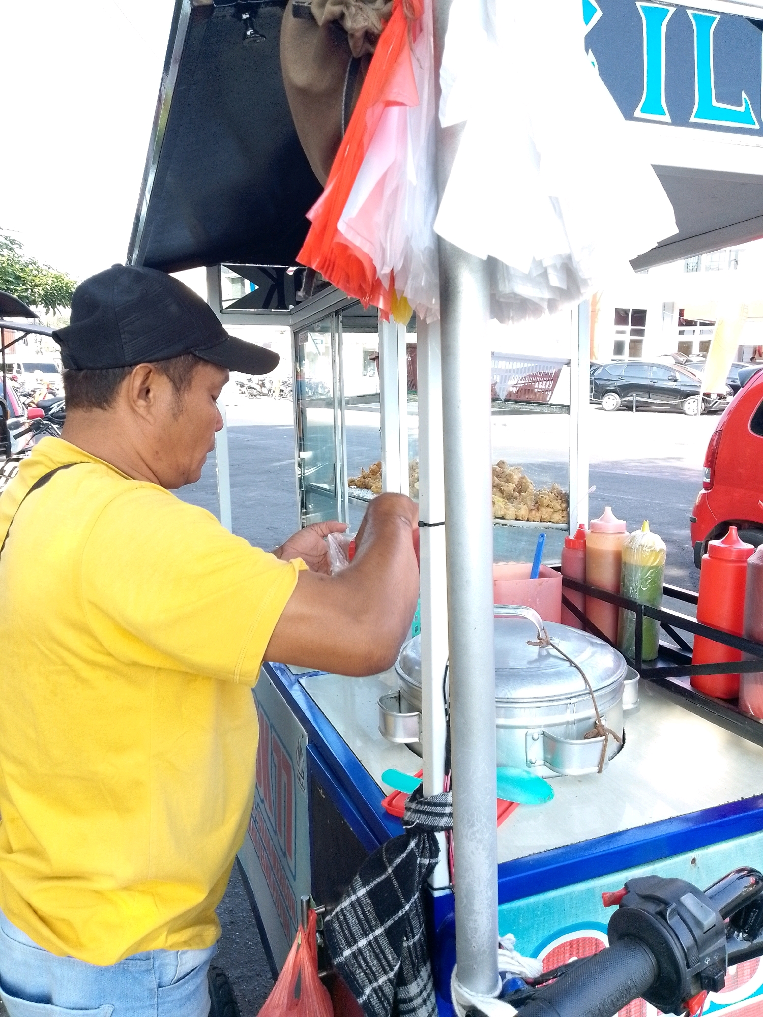 A vendor, a man wearing a black baseball cap and a yellow t-shirt, stands behind a food cart. He is holding a clear plastic bag in his right hand and appears to be preparing food. The food cart has a metal frame and a blue and white exterior. Several condiment bottles are visible on the cart, as are a metal pot and a blue scoop. In the background, there are parked cars and motorcycles. The scene appears to be an outdoor market or street.