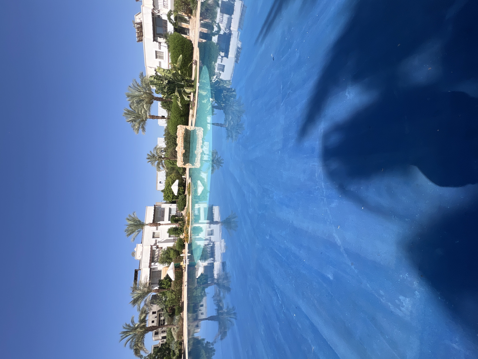 The image depicts a bright, sunny day at a resort in Sharm El Sheikh, Egypt, with a large, vibrant blue swimming pool dominating the foreground. The pool's surface reflects the clear blue sky and the surrounding white buildings, lush greenery, and palm trees. In the background, white, modern resort buildings with balconies and shaded areas are visible, interspersed with tropical plants and palm trees. A decorative rock formation with water cascading over it is situated within the pool area, adding to the resort's ambiance. The overall scene conveys a sense of tranquility and vacation, characteristic of a resort destination.