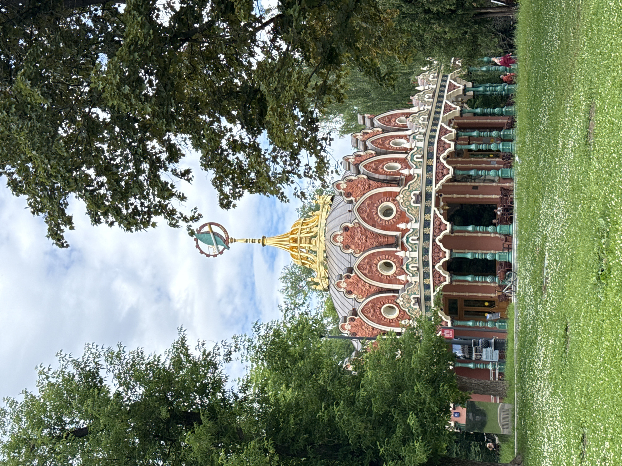 The image captures the picturesque "Pavilion of Ukraine" at VDNKh in Moscow, Russia. This ornate building, with its distinctive red and gold facade, features a domed roof topped by a globe-like ornament. The pavilion is surrounded by lush greenery and a grassy lawn dotted with small white flowers. Several trees frame the scene, with their leaves creating a dappled effect of light and shadow. In front of the pavilion, there's a covered walkway supported by decorative green columns. A bicycle is parked near the entrance, and a few patrons are visible sitting at tables outside the pavilion. In the foreground, the vibrant green lawn stretches out, inviting a sense of tranquility and leisure. The sky above is partly cloudy, with a mix of blue sky and white clouds, suggesting a pleasant day. A few individuals are seen walking on the grass in the background, adding a human element to the serene landscape.