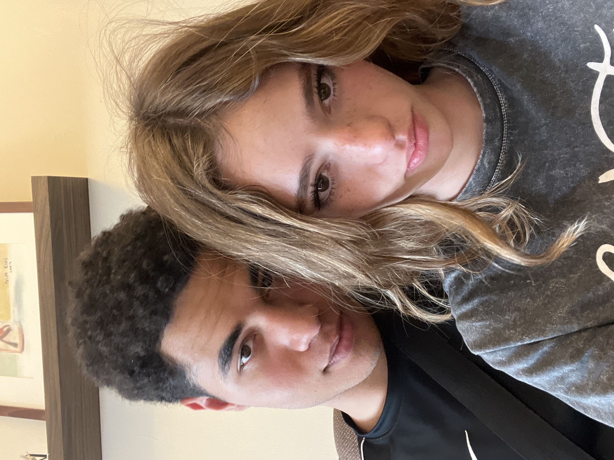 Two individuals, a female with light brown hair and a fair complexion, and a male with dark curly hair and a tan complexion, are positioned closely together. The female is wearing a gray top, while the male is wearing a black shirt with a white logo. The background includes a beige wall and a framed picture. The image does not provide information about San Bernardino Tlaxcalancingo, Mexico, or the city.