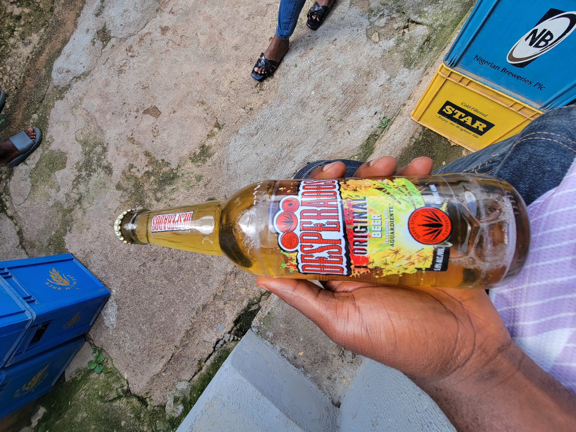 This media file shows a close-up of a person's hand holding a bottle of Desperados Original Beer. The bottle is mostly full of golden-colored liquid and has a label with red and yellow text that reads "DESPERADOS ORIGINAL BEER FLAVOURED WITH AGUARDIENTE 5.9% ALC/VOL". In the background, there are several blue and yellow plastic crates, one of which has "NB Nigerian Breweries Plc" printed on it. Another crate has a yellow "STAR" logo with the text "...Enjoy the Brighter life". The ground appears to be concrete or a similar paved surface, with some patches of moss or dirt. The scene suggests an outdoor setting, possibly a casual gathering or a break during an event, with the presence of beer crates indicating a place where drinks are served. The lighting suggests it could be daytime. The overall mood conveyed is relaxed and informal.