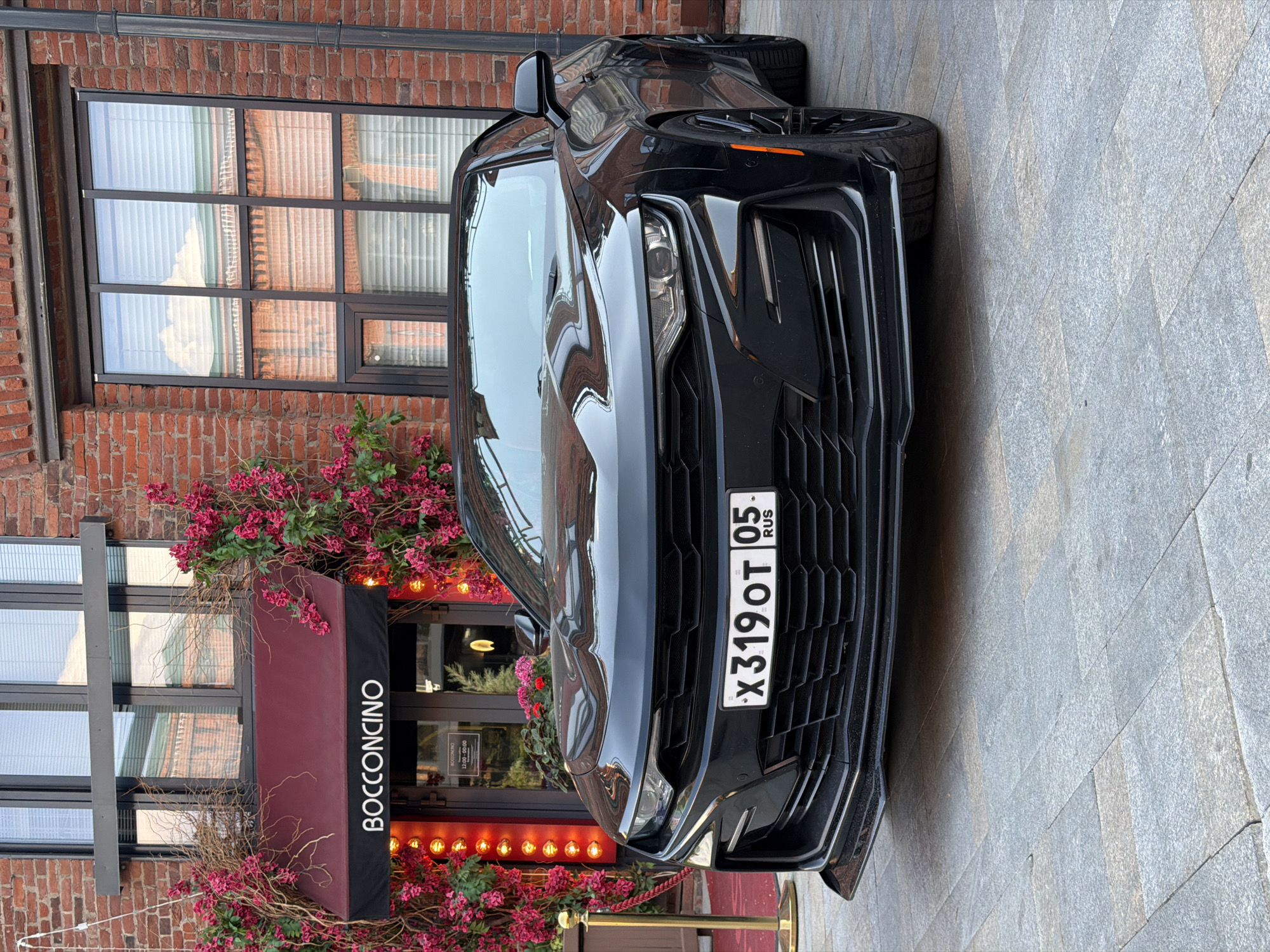 The image captures a black Chevrolet Camaro parked in front of the Bocconcino restaurant, as seen in the lower left. The facade of the building is brick, with a window above the restaurant entrance. Red floral decorations surround the entrance and window. The vehicle has a Russian license plate, "X319OT 05 RUS," indicating it is registered in Russia. The setting appears to be urban, and based on the restaurant name and license plate, it is likely Moscow, Russia. The scene appears to be an urban environment.