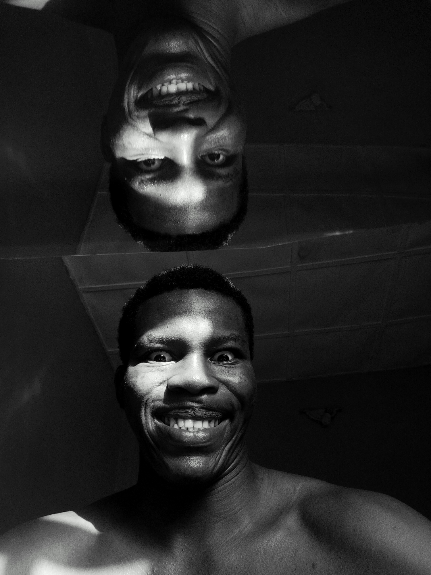 The image is a black and white portrait of a dark-skinned man, with his face occupying the lower half of the frame. Above him, there is an upside-down reflection of his face. The man is smiling broadly, showing his teeth. A strong light source illuminates parts of his face, creating stark contrasts and shadows. The background appears to be a ceiling with a rectangular grid pattern. The location is potentially in Keffi, Nigeria, as the image context indicates, although there are no specific objects in the image that pinpoint the location. The man is shirtless, indicating an indoor environment.