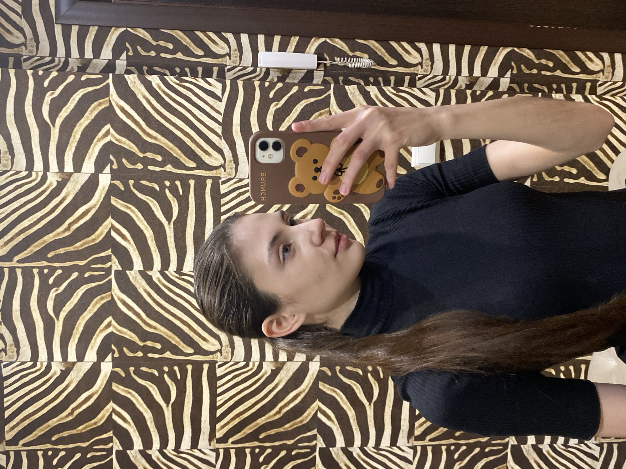 A person is lying on a surface with a zebra-striped pattern. They are holding a brown phone with a cartoon bear on the case and taking a selfie. The person has long brown hair and is wearing a black top. In the upper portion of the image, there is a dark brown beam above the zebra-striped surface and some electronic equipment.
There is no discernible information that indicates this image was taken in Votkinsk, Russia.