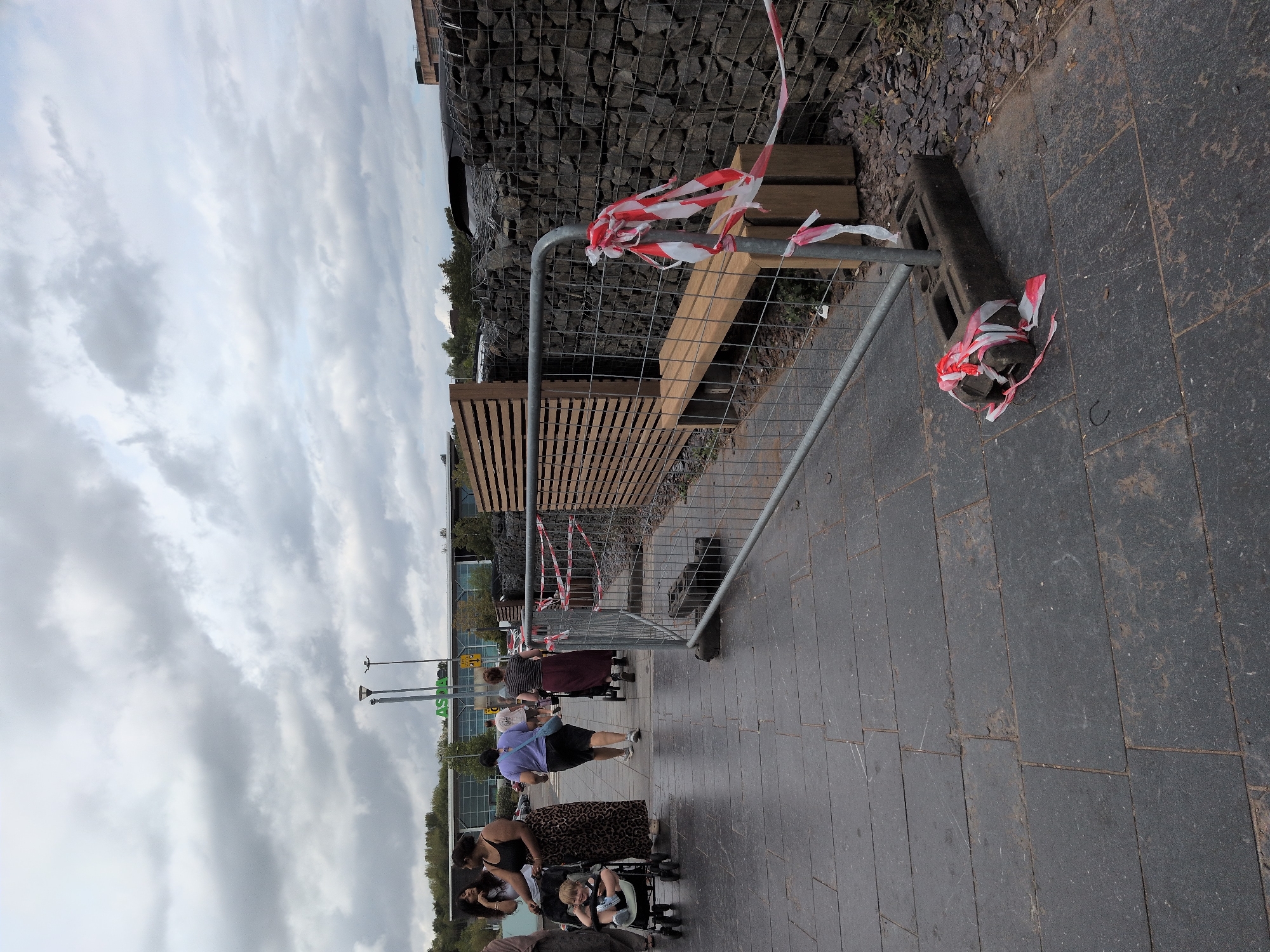 In Telford, United Kingdom, on a cloudy day, a temporary metal barrier with red and white tape cordons off an area next to a gabion wall. A wooden bench is visible behind the barrier. Several people are walking on a paved walkway in front of the barrier. One person is pushing a stroller with a child in it, and another person is pushing a shopping cart. In the background, the Asda logo is visible on a building.