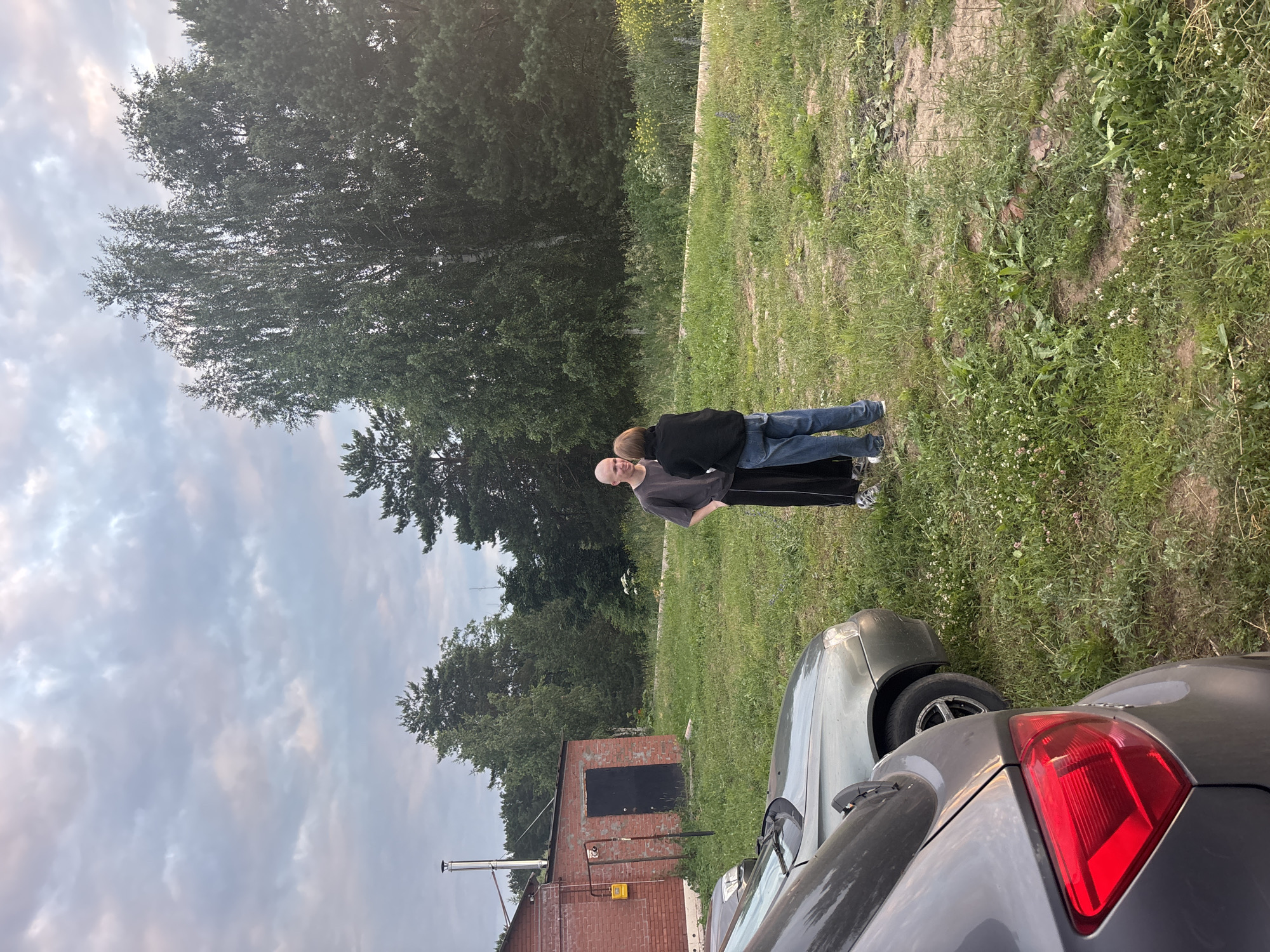The image depicts an outdoor scene with two individuals standing on a grassy area. One person, appearing to be a man with a shaved head, is wearing a dark t-shirt and dark pants with a white stripe. He is facing towards another individual, who is wearing a black jacket and blue jeans. They appear to be in conversation or interacting with each other. In the foreground, the rear of a grey car is visible, and a portion of a silver car is also present. Behind the individuals, there is a brick building with a black door and a chimney. The background consists of trees and a cloudy sky, suggesting a temperate climate. The scene is likely in a rural or semi-rural area, possibly a dacha or a backyard. The location is specified as Chaikovsky, Russia.