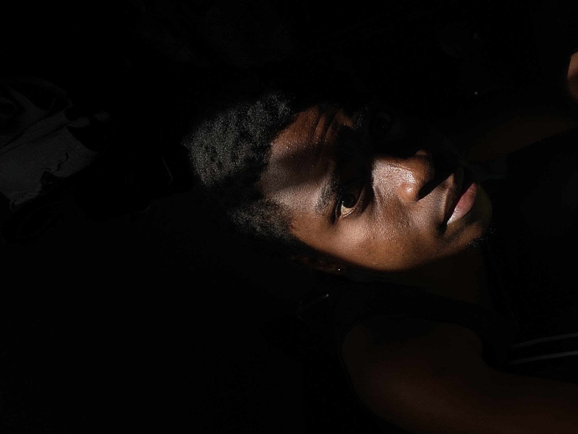 A low-angle close-up photograph shows a man with dark skin partially illuminated by a light source that creates strong contrasts. The light highlights his forehead, nose, right eye, and mouth, while the rest of his face and body are obscured in deep shadow. His hair is short and dark, and there's a hint of stubble on his chin. A piece of clothing is visible in the upper left corner, also cast in shadow. The photograph has a somber and dramatic mood due to the high contrast and limited visibility. There is no indication of a location in the city of Onitsha, Nigeria.