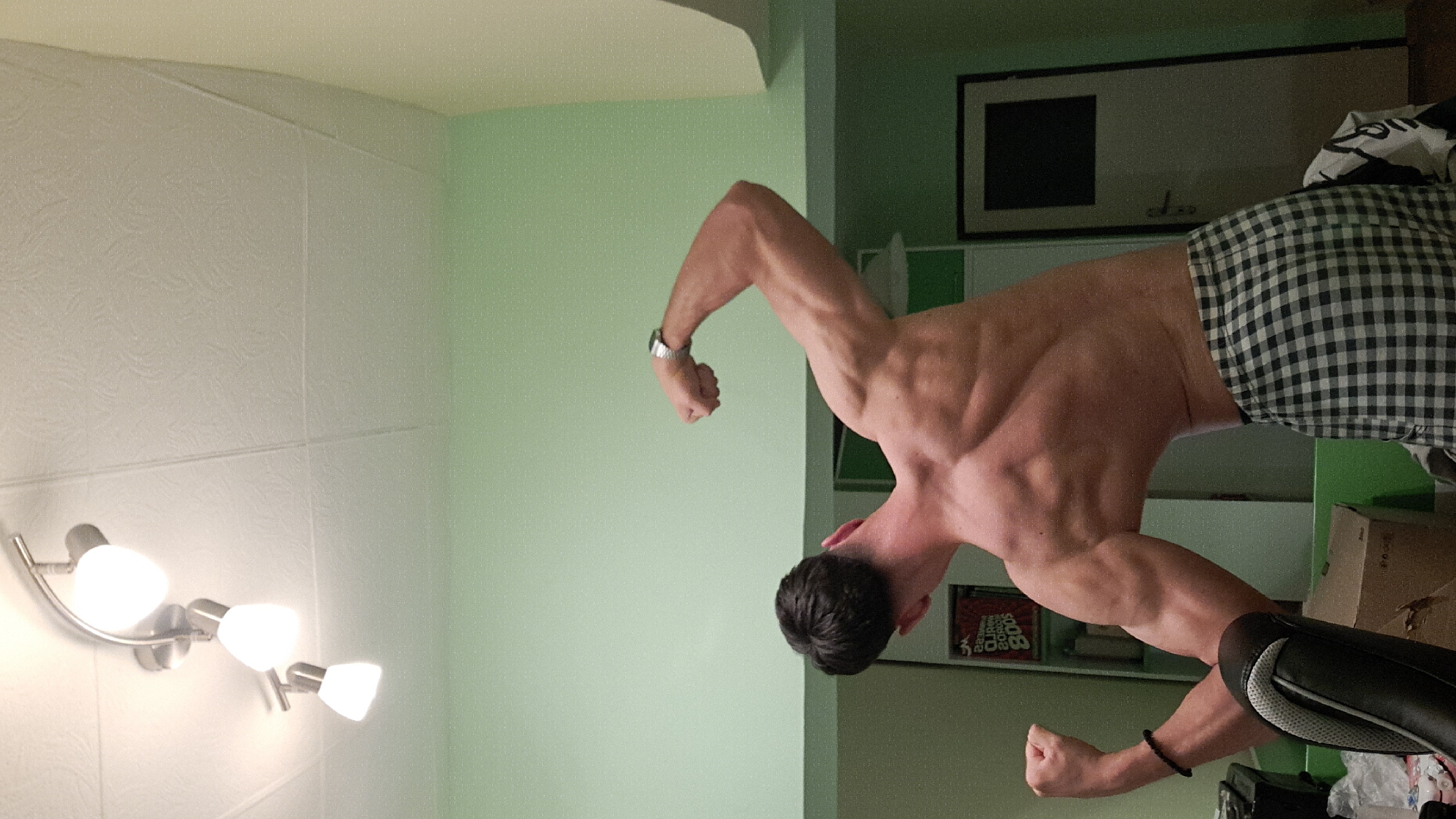 A muscular man, shirtless and wearing checkered shorts, is posing with flexed biceps. He is facing away from the camera. A watch is visible on his left wrist and a bracelet on his right. The setting appears to be indoors, with a light fixture on the left wall. The wall behind the man is painted green. A shelf containing books and a framed picture are visible in the background. A chair is visible in the foreground. There is no specific information to infer that the image was taken in the city of Burgas, Bulgaria.