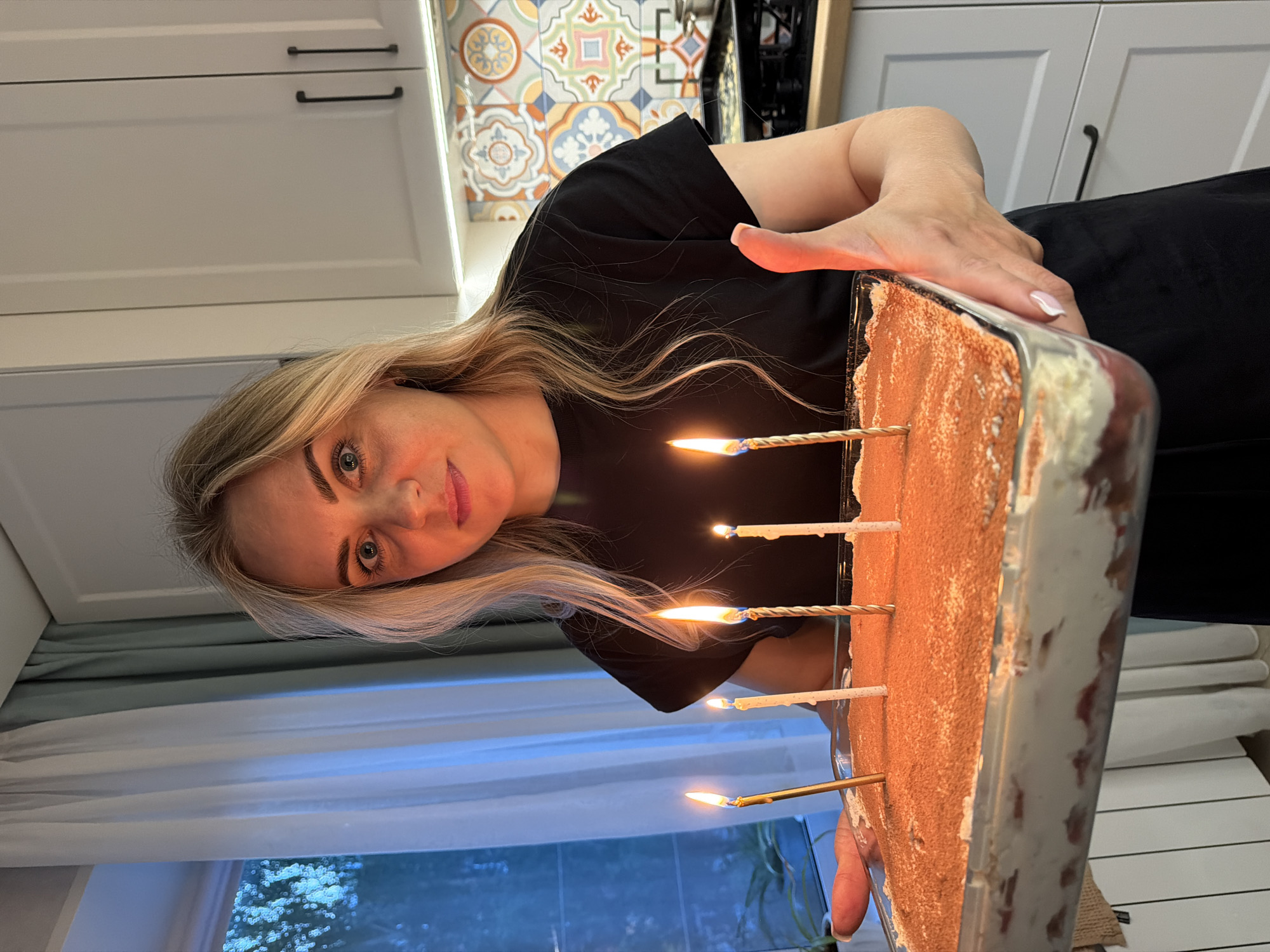 A woman is holding a rectangular cake with several lit candles in it. The woman has blonde hair and is wearing a black shirt. The cake appears to be dusted with a brown powder, possibly cocoa, and has a light-colored edge. The background shows a kitchen setting with white cabinets and decorative tile backsplash. Outside the window, greenery is visible. The location is likely indoors and cannot be definitively confirmed to be in Votkinsk, Russia based solely on the image.