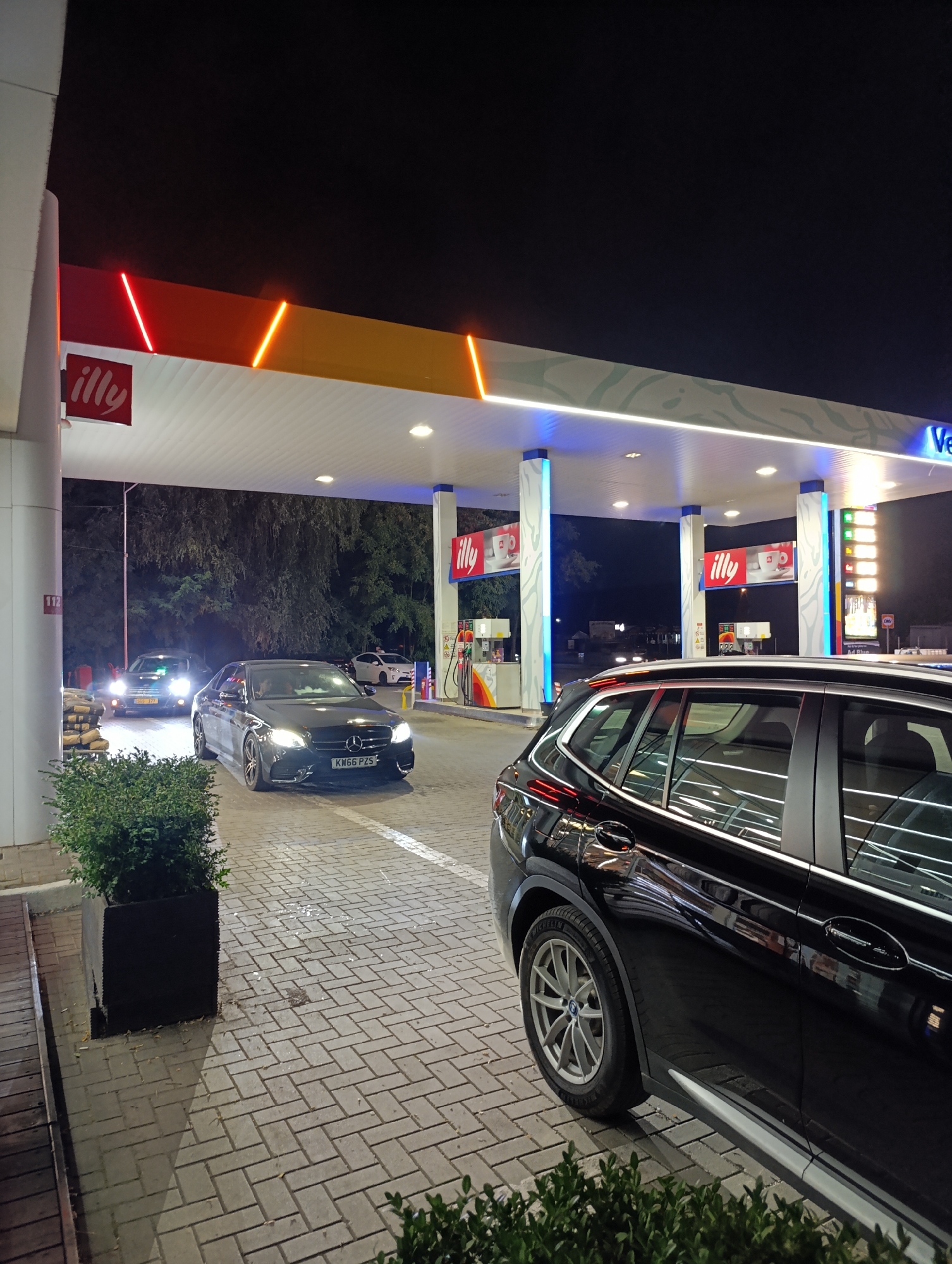 The image depicts a night scene at a Shell gas station located in Colonita, Moldova. In the foreground, a black SUV is parked on the right side of the frame, partially obscuring a black Mercedes-Benz sedan in the middle ground, which has its headlights on. Another car, a dark sedan with its headlights illuminated, is visible further back on the left. The gas station canopy is illuminated by red, orange, and blue lights. "Illy" logos are prominently displayed on the canopy and on signs. In the background, trees and the night sky are visible. The ground is paved with interlocking brick pavers.